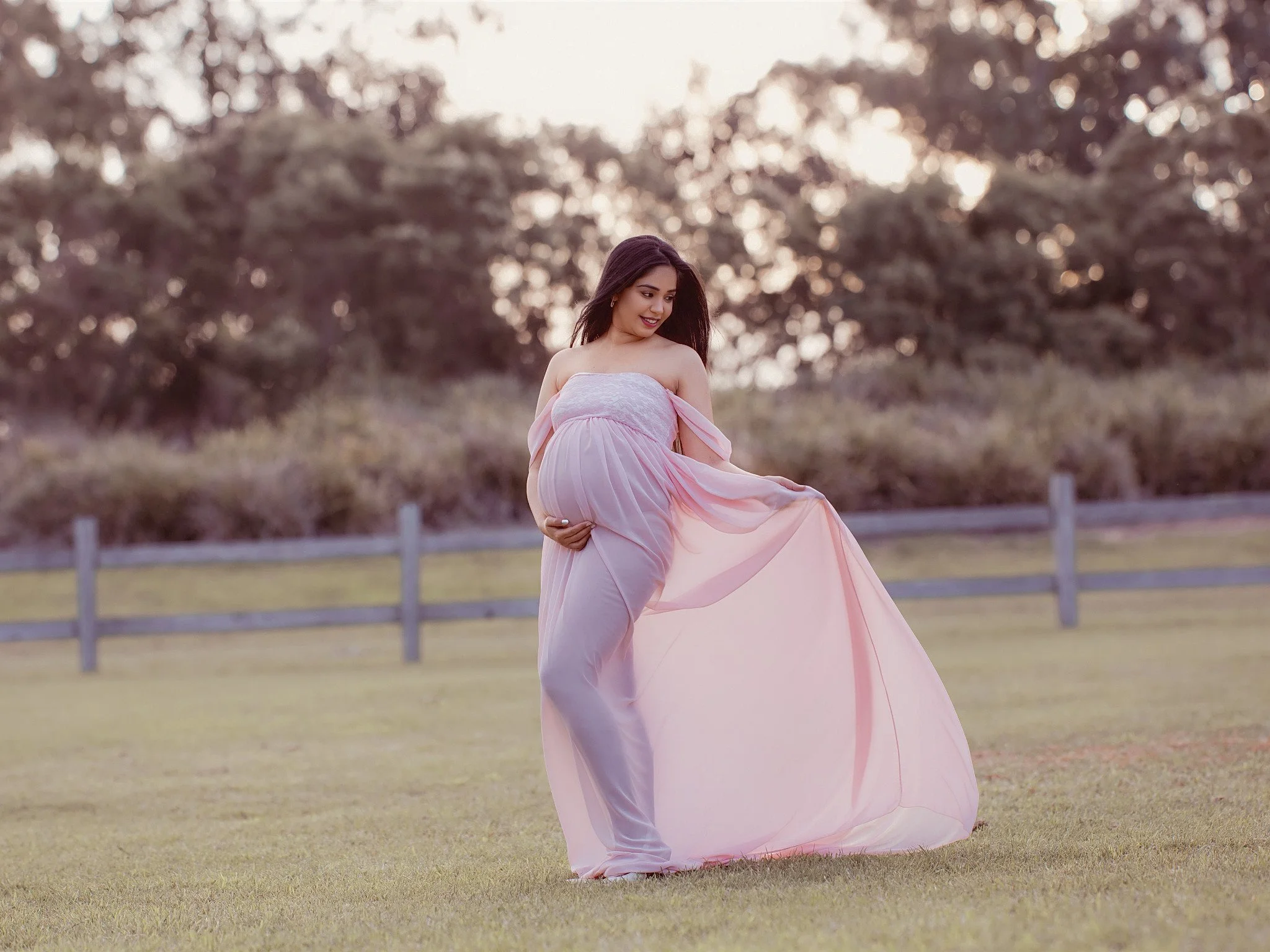 A pregnant woman in a flowing pink dress stands on a grassy field with trees in the background, holding her belly and looking down at it.