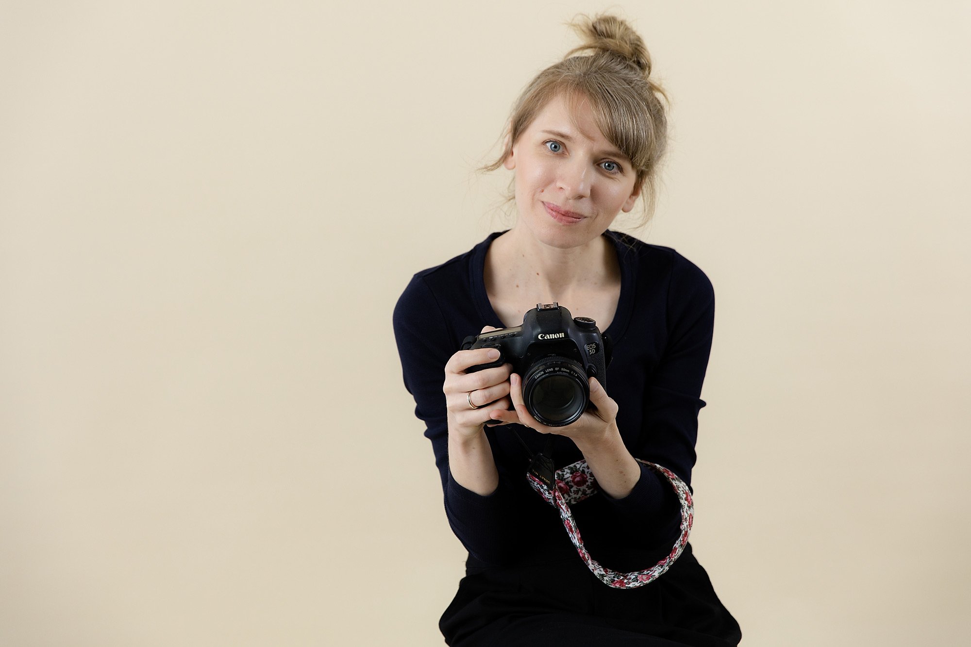 A woman with blonde hair in a bun holding a Canon DSLR camera and looking at the camera.