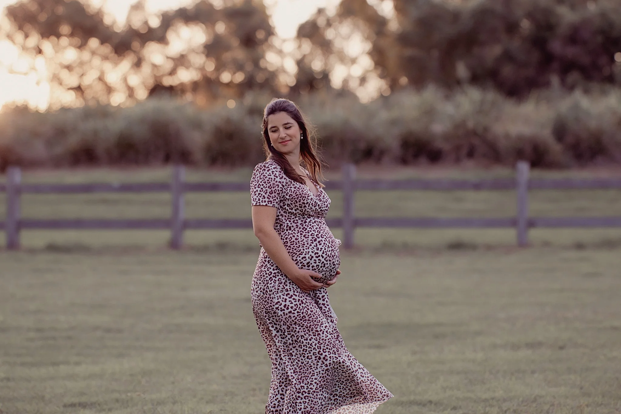 Pregnant woman in a leopard print dress standing outdoors in a grassy field, holding her belly, with a wooden fence and trees in the background during sunset.