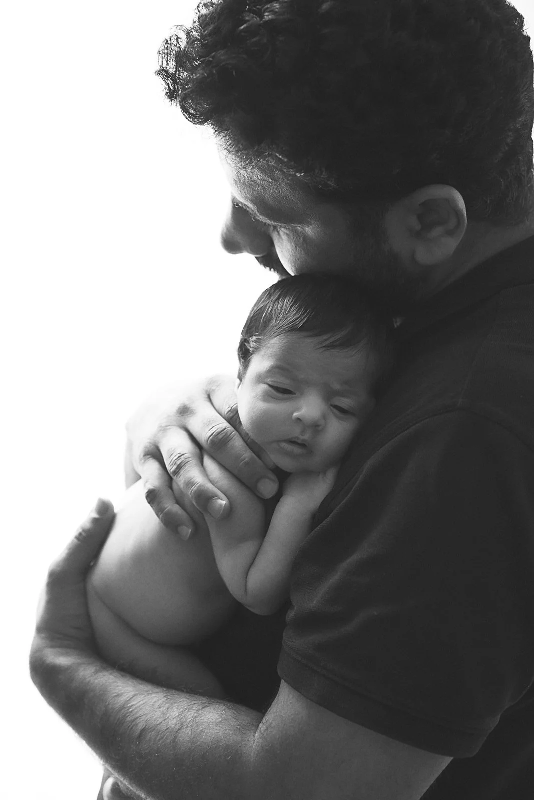 A man holds a newborn baby close, gently cradling the baby's head and shoulder with both hands. The man is leaning in, touching his forehead to the baby's head, and both are viewed in profile against a plain background.