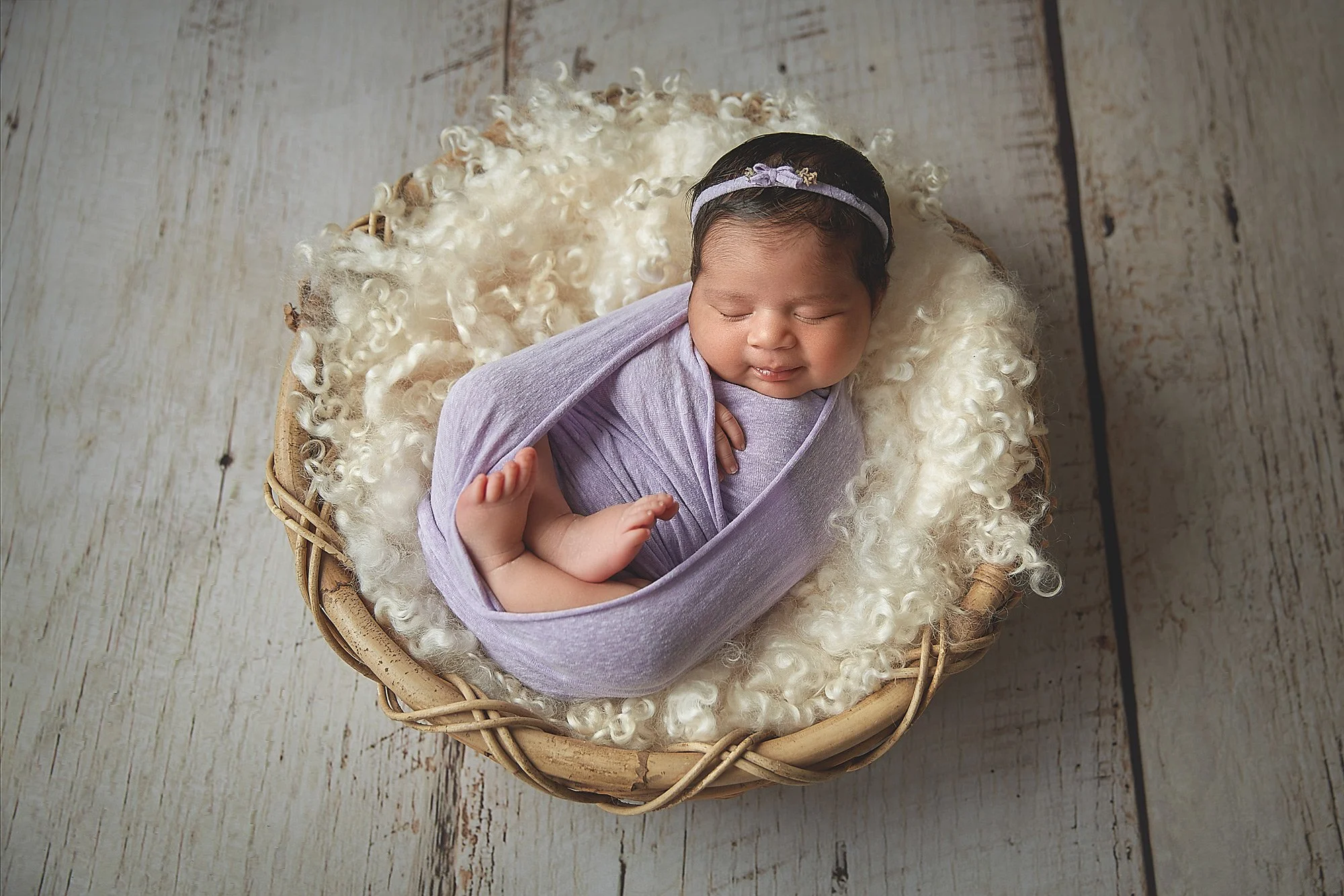 A peaceful newborn baby wrapped in a lavender swaddle, sleeping in a wicker basket lined with soft, curly white fur, on a light wooden floor.