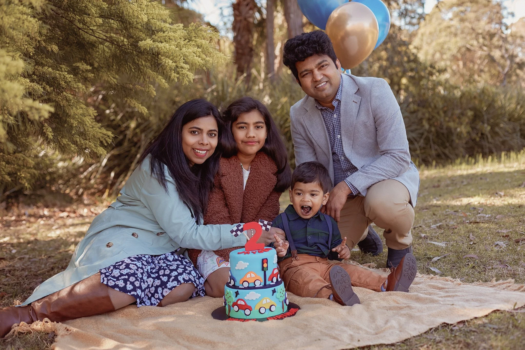 A family of four celebrating a child's second birthday outdoors, sitting on a beige blanket with a colorful car-themed birthday cake and balloons in the background.