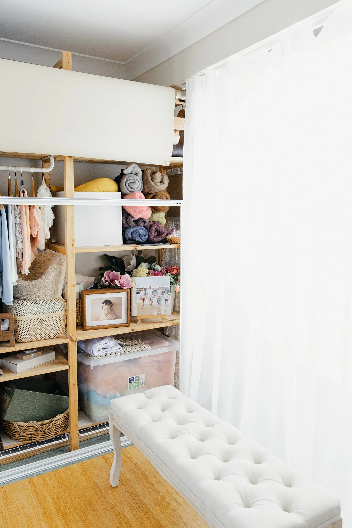 A white tufted bench with cabriole legs in front of a wooden shelving unit filled with folded blankets, photos, and household items, next to a partially visible window with white curtains and a hardwood floor.