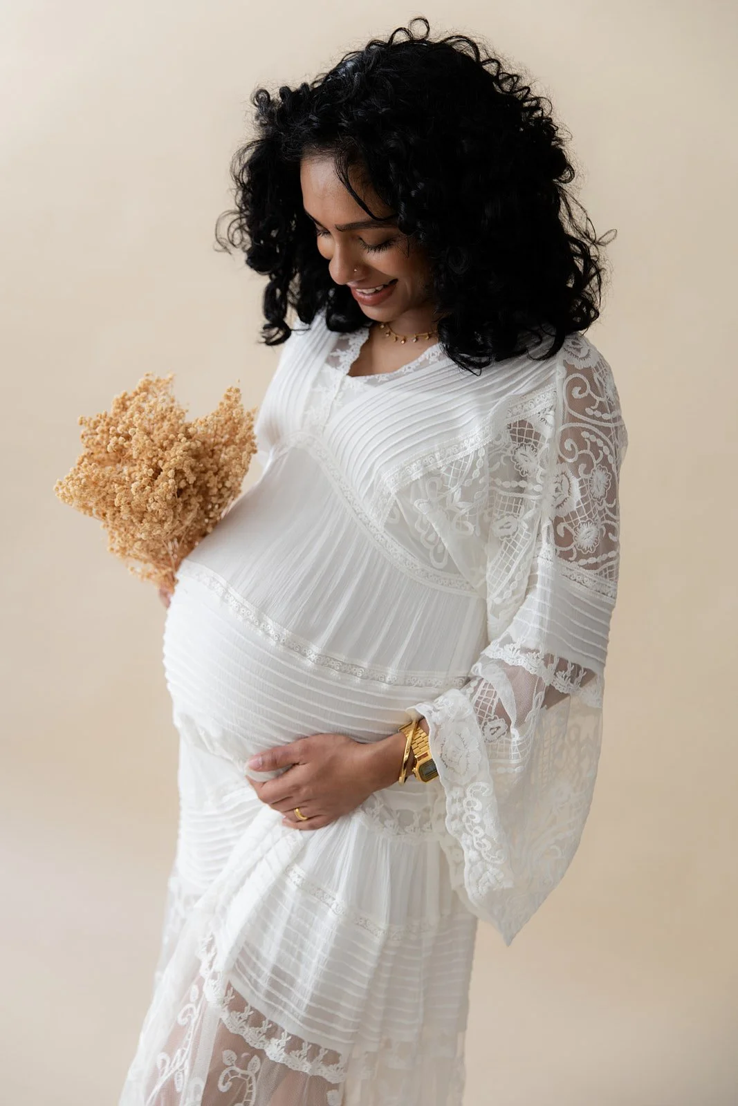 Pregnant woman in a white lace dress holding a bouquet of dried flowers, smiling gently, against a plain beige background.