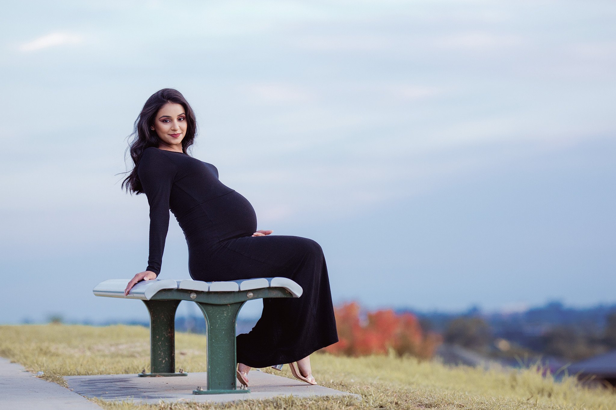 Pregnant woman in a long black dress sitting on a park bench outdoors during daytime.