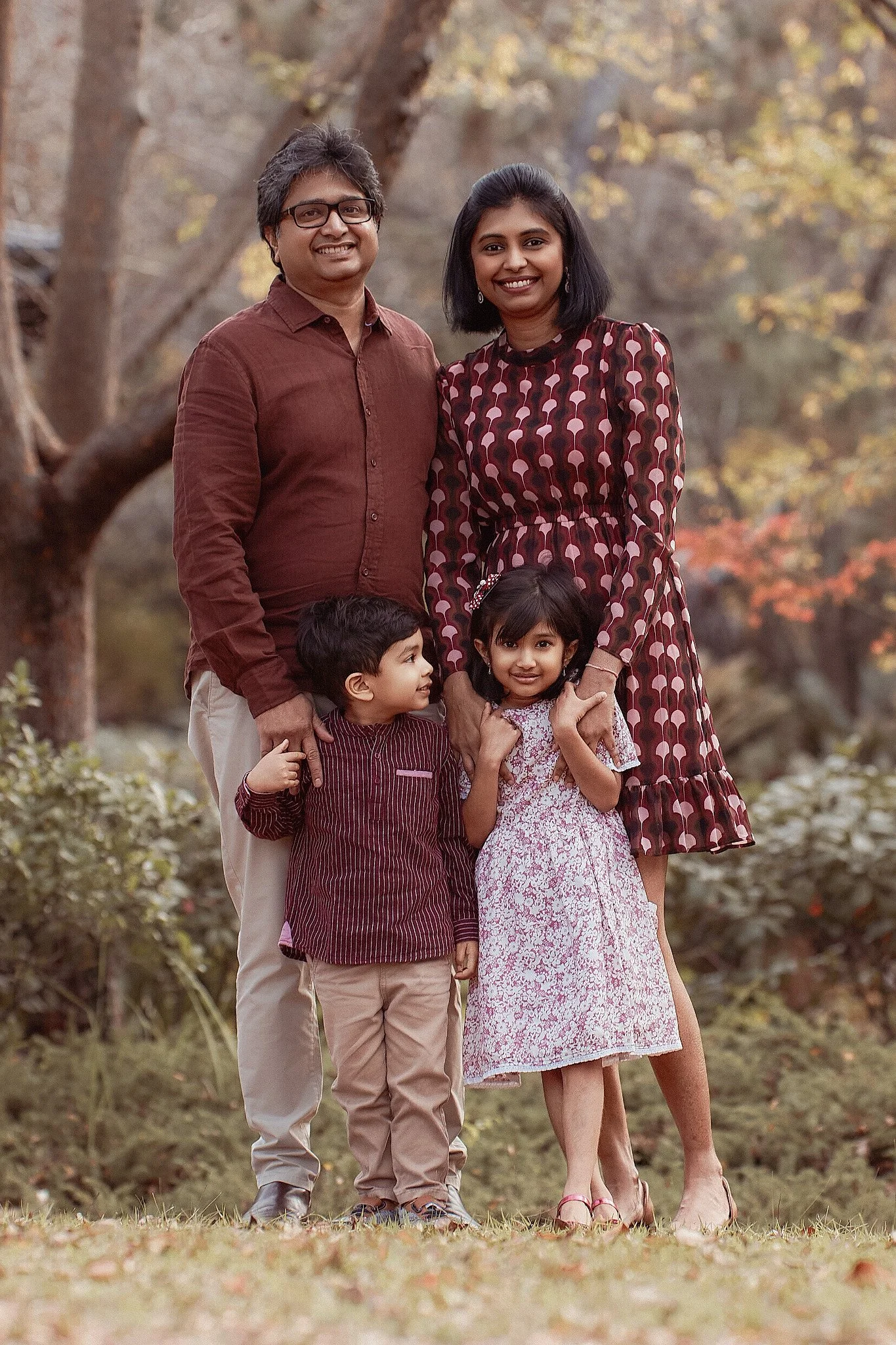 A smiling family of four outdoors in a park with autumn trees, including a man, a woman, a young boy, and a young girl.