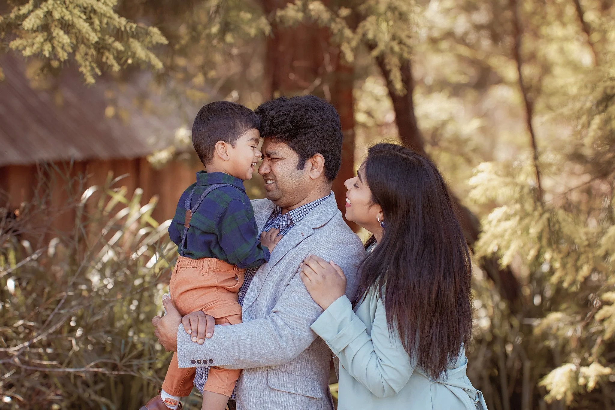 Family enjoying outdoor moment, father holding young son, mother smiling, in a park with trees and sunlight in the background.