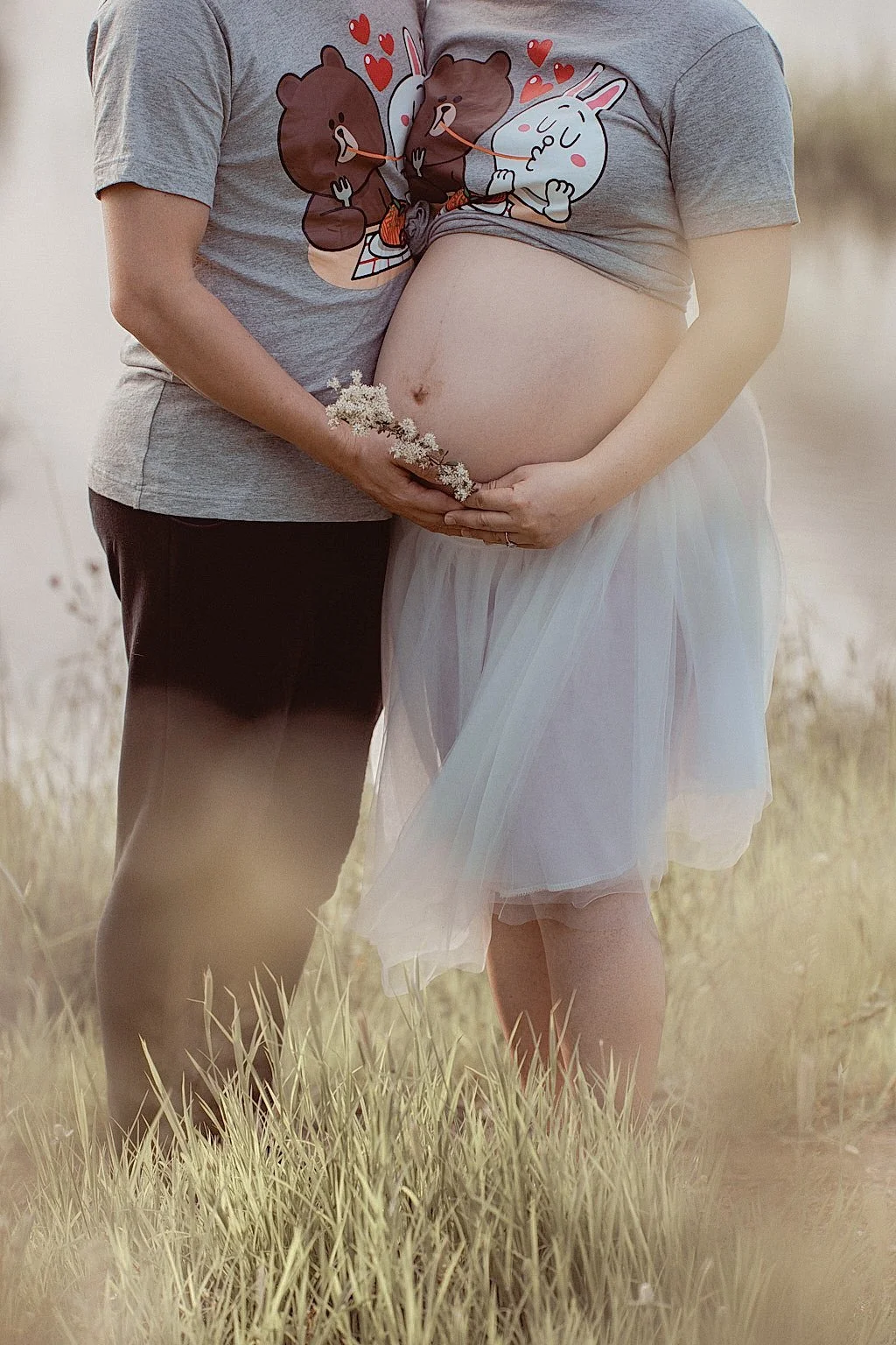 Pregnant woman and partner standing in a field, holding her belly, with woman wearing a light tulle skirt and man in casual clothes, both holding a flower, with cartoon bear and bunny on her shirt.
