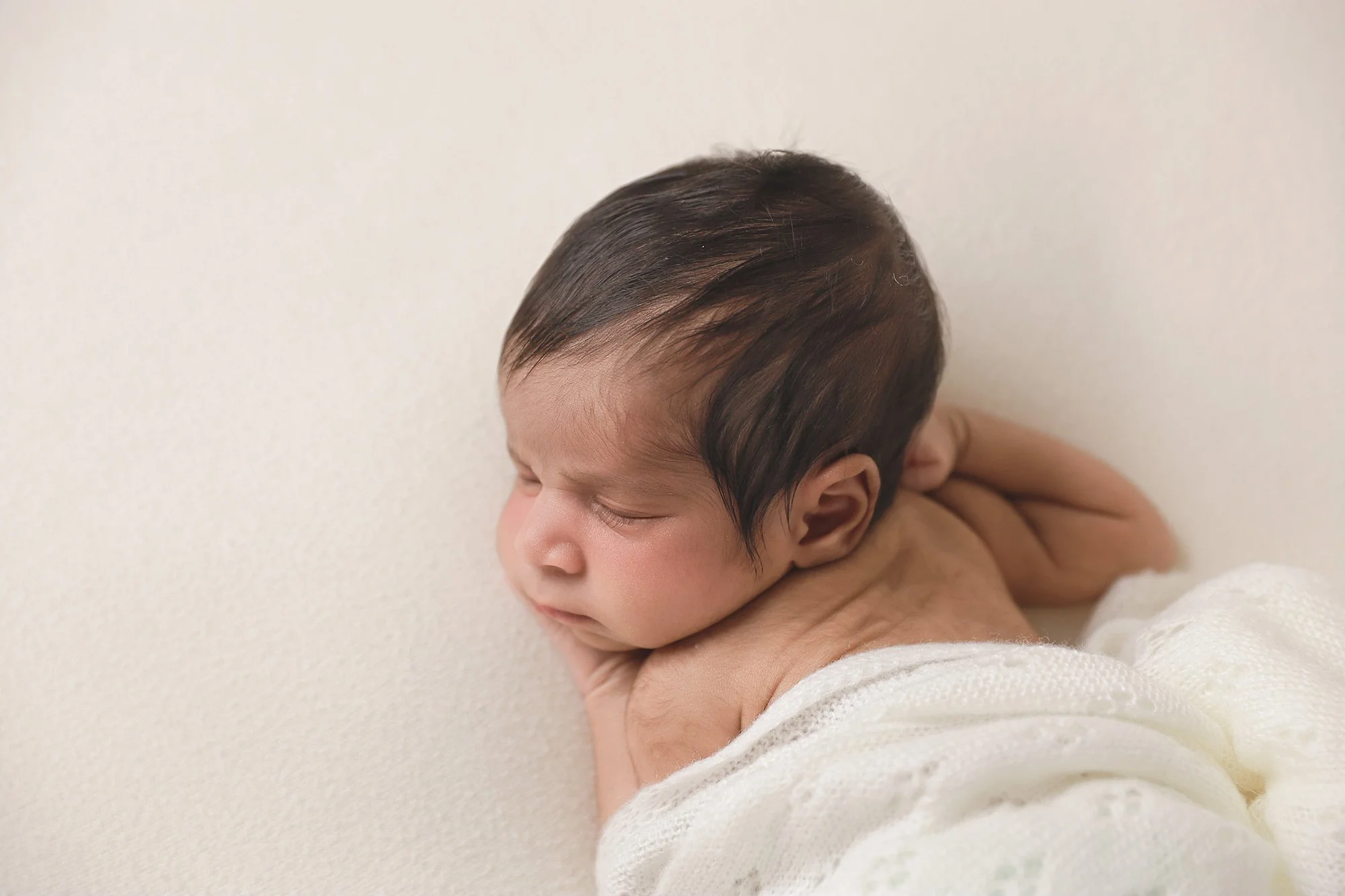 Close-up of a newborn baby with dark hair, resting face down on a soft, cream-colored surface, wrapped in a white blanket.