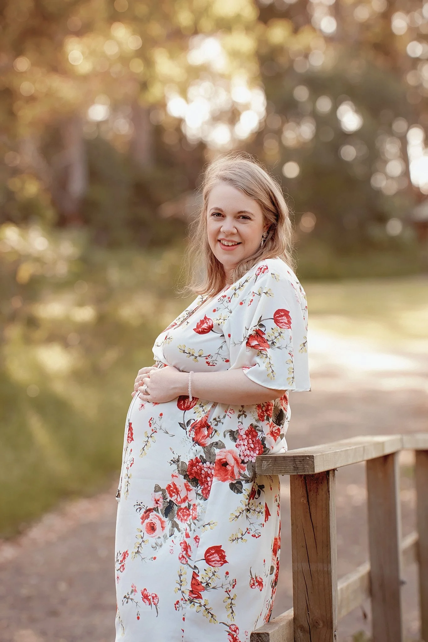 A pregnant woman with blonde hair wearing a white dress with red floral print, standing outdoors beside a wooden railing near a body of water, smiling at the camera with trees and soft sunlight in the background.
