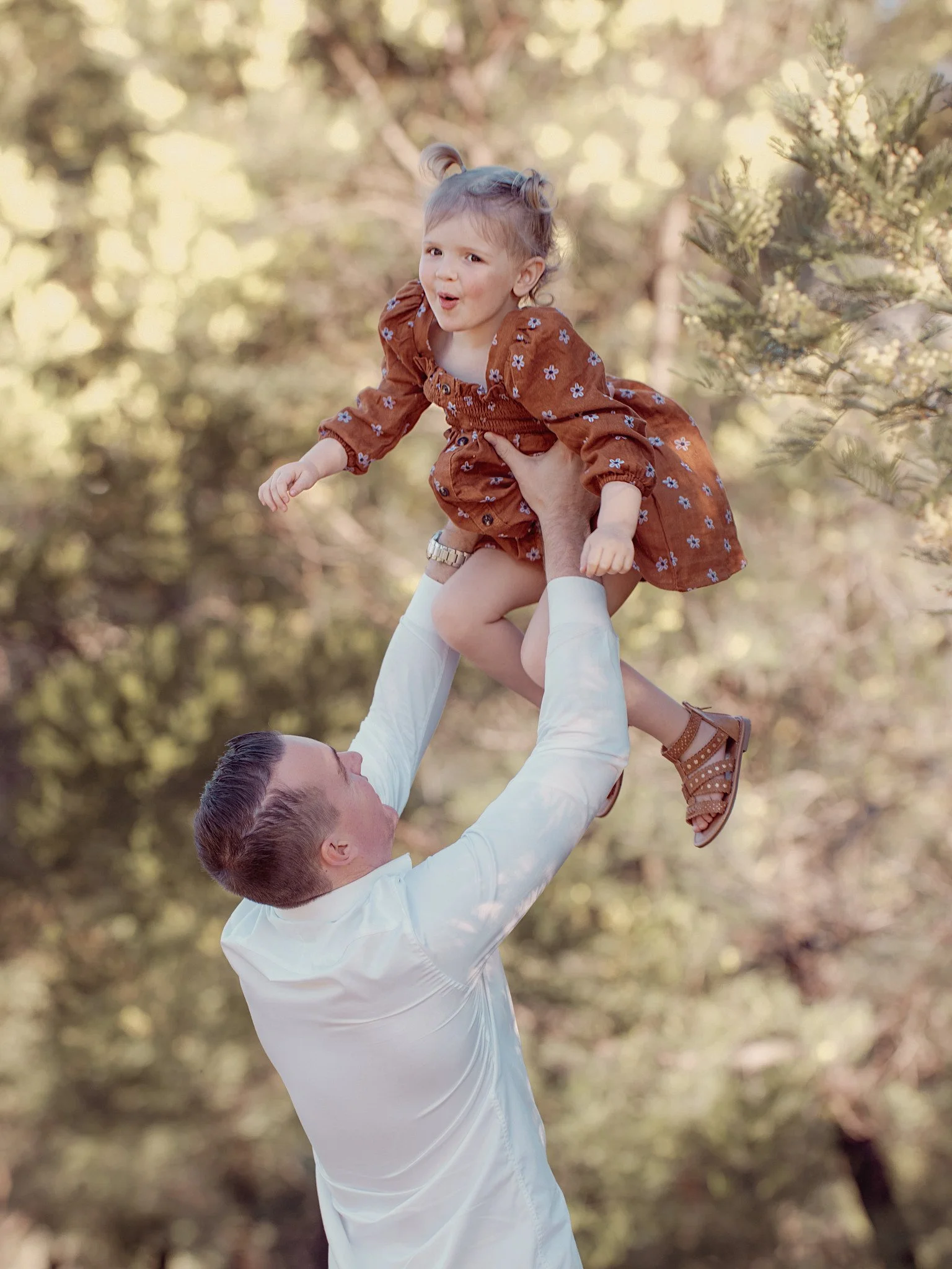A man lifting a young girl in the air outdoors surrounded by trees.