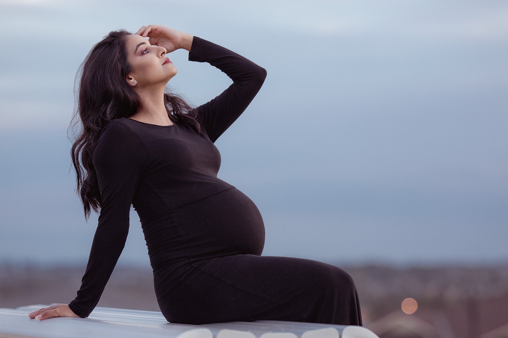 A pregnant woman in a black dress sitting outdoors on a white surface, looking up with her hand on her forehead, against a cloudy sky background.
