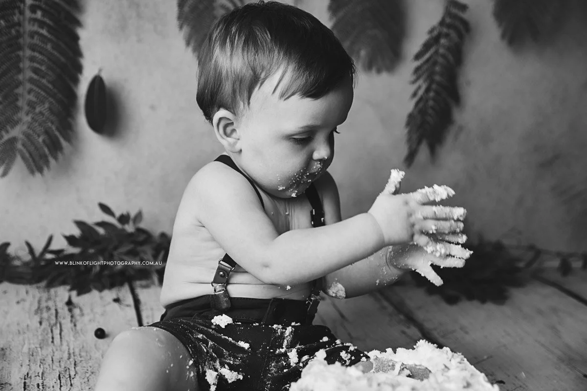 A young child with short hair sitting on the floor, covered in flour or dough, playing with it and using both hands to explore, with a backdrop of plants and a textured wall.