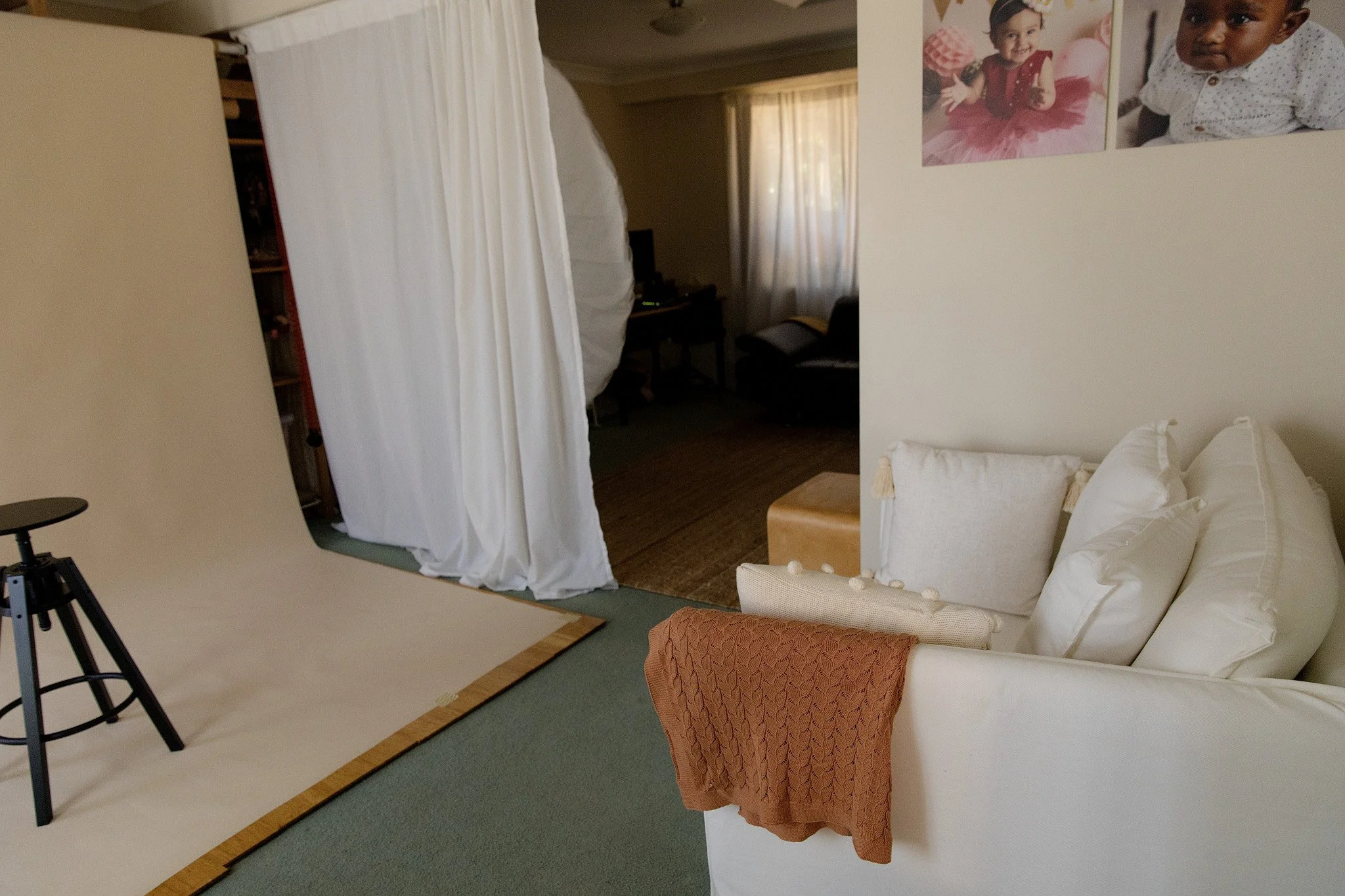 An indoor room with a beige wall and a large white sheet or backdrop set up on a wooden platform. There is a tripod visible in the foreground. The room contains a couch with a reddish-orange patterned blanket draped over the armrest. The wall has two large photos of a young girl, one smiling and the other looking serious, both in casual clothing. In the background, there are curtains on window and some furniture including a desk with a computer monitor.