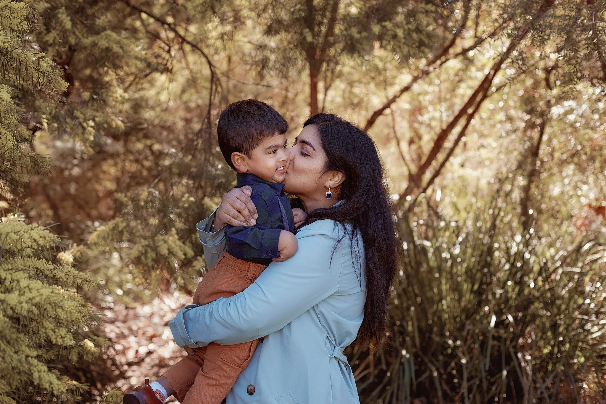 A woman holding a young boy and giving him a kiss on the cheek outdoors surrounded by trees and foliage.