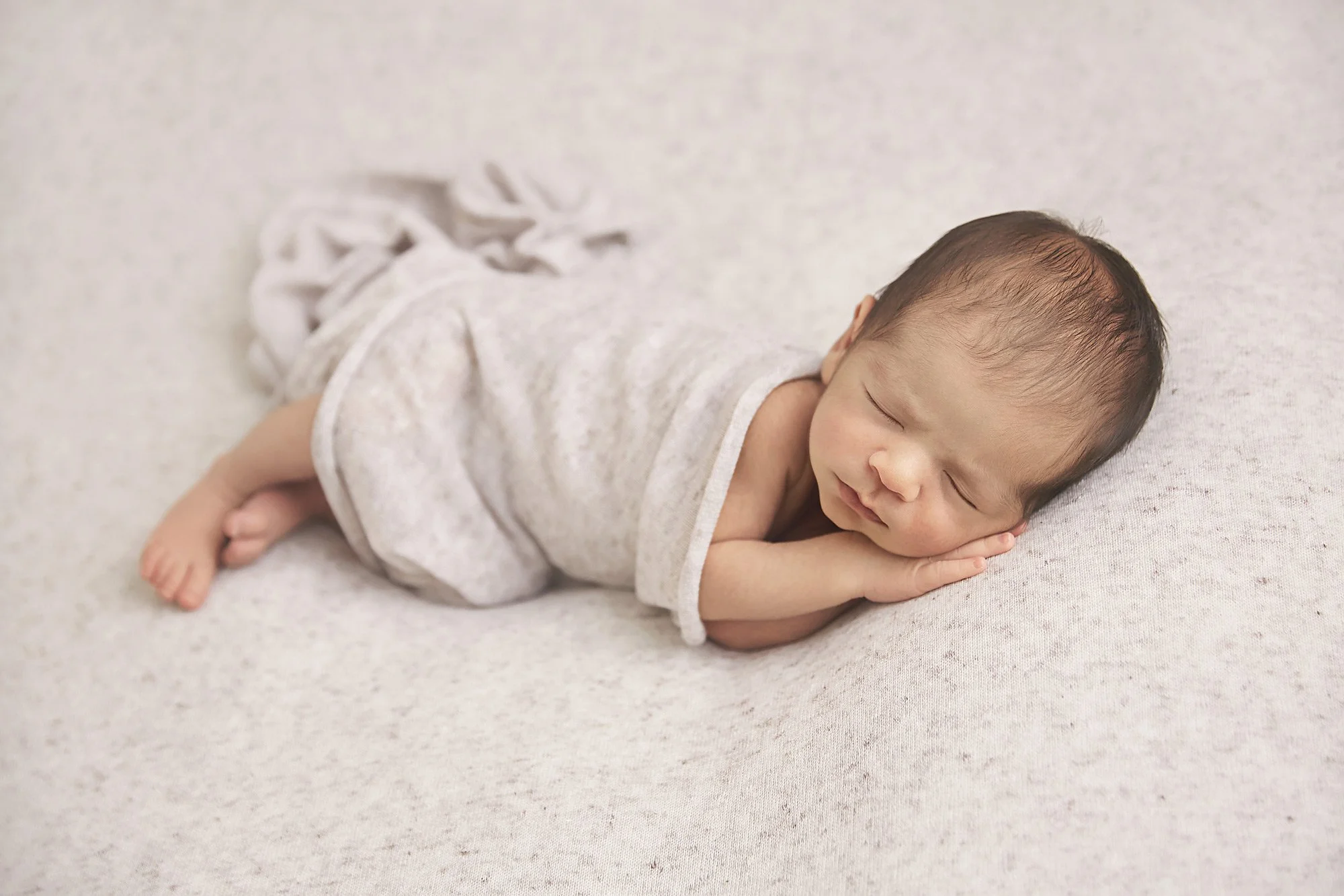 A sleeping baby lying on their side on a light-colored surface, dressed in a light gray outfit, with a peaceful expression, resting their head on their hand.
