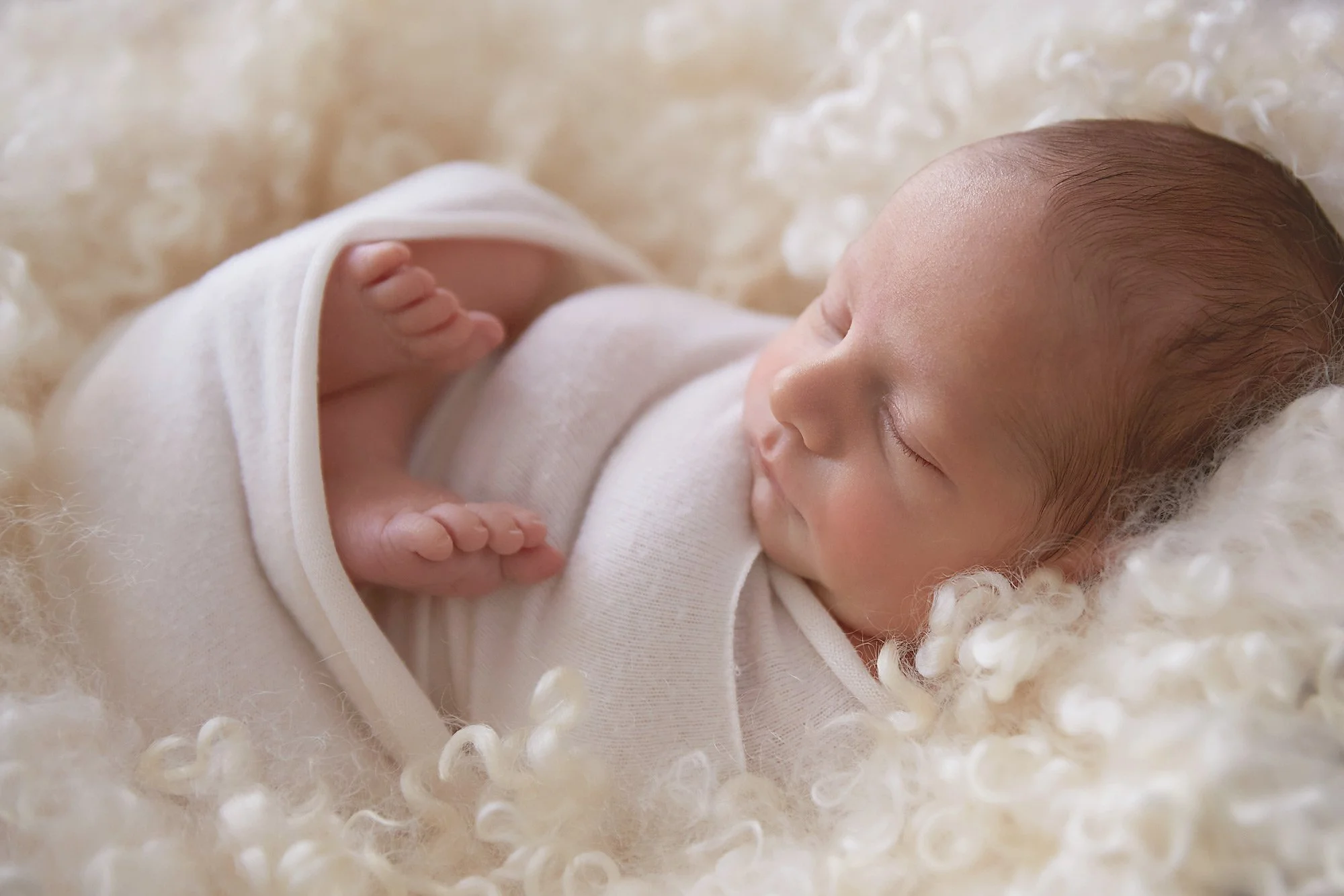 A sleeping baby lies on a soft, fluffy cream-colored blanket, wearing a white outfit, with hands gently clasped near the face.