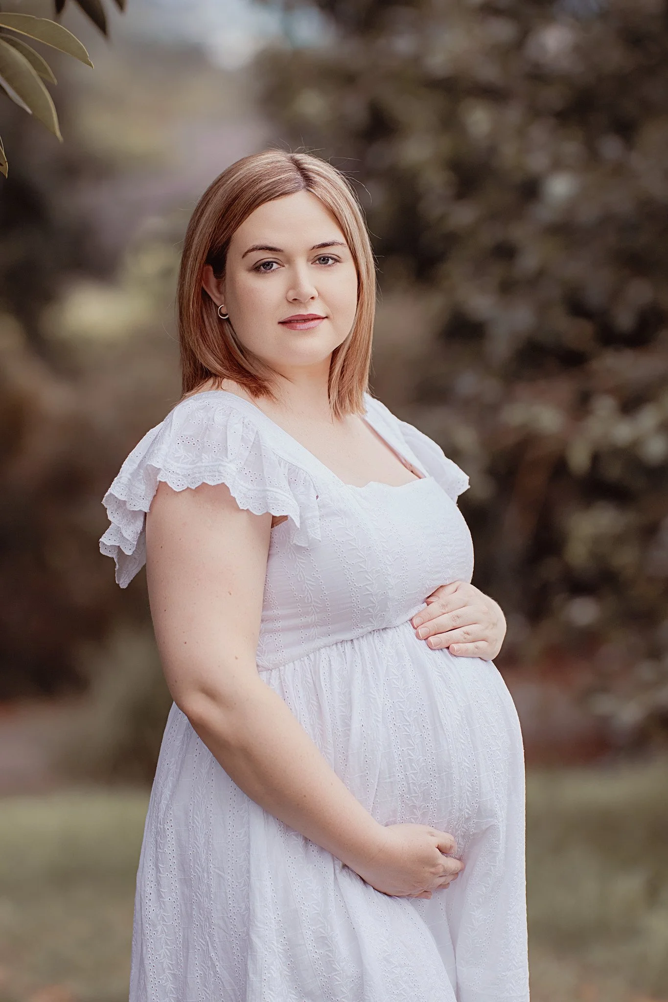 A portrait of a pregnant woman with shoulder-length brown hair, wearing a white dress with ruffled sleeves, standing outdoors with blurred trees in the background.