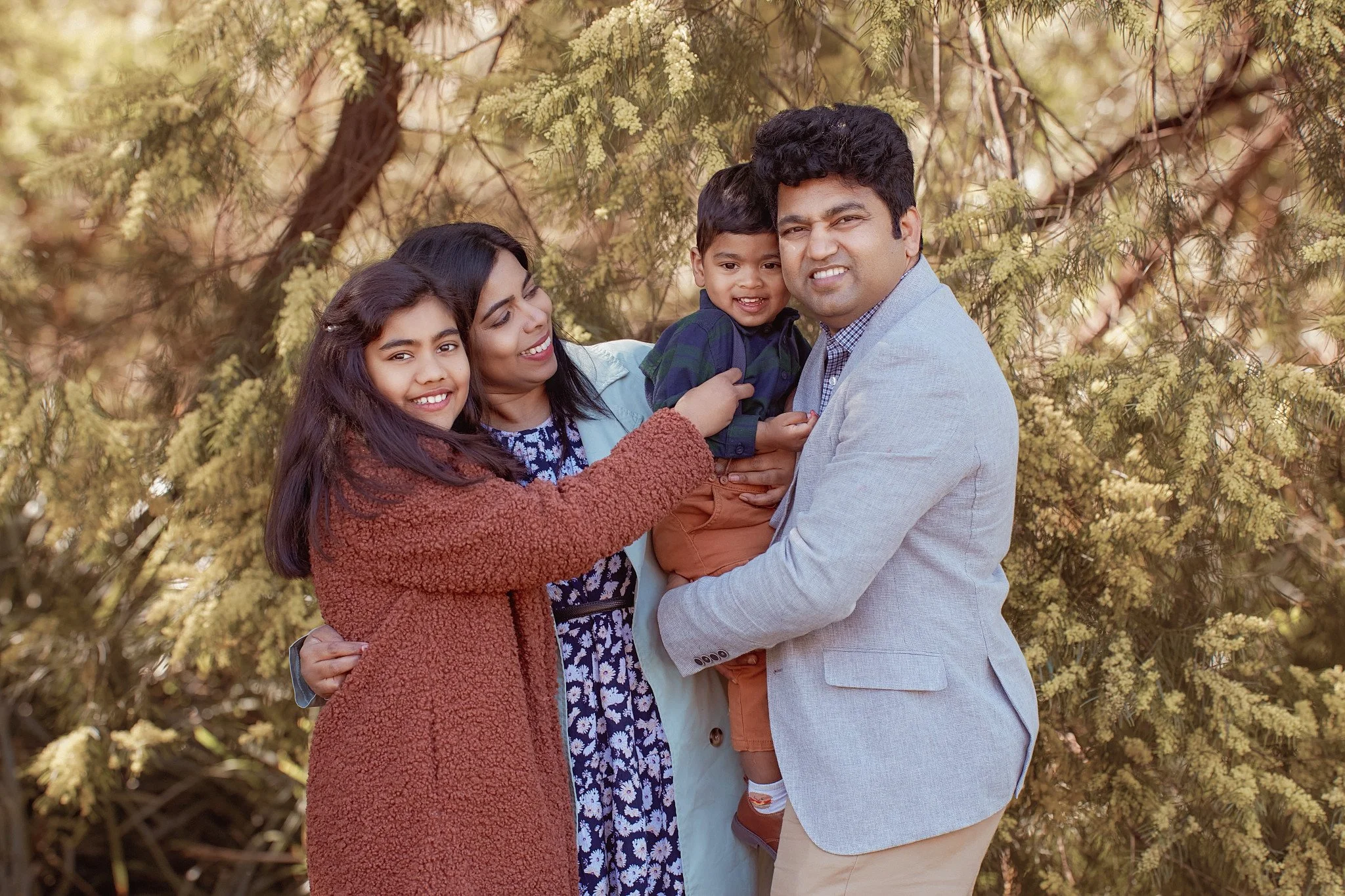 A family of four outdoors in front of green foliage, smiling and hugging each other.