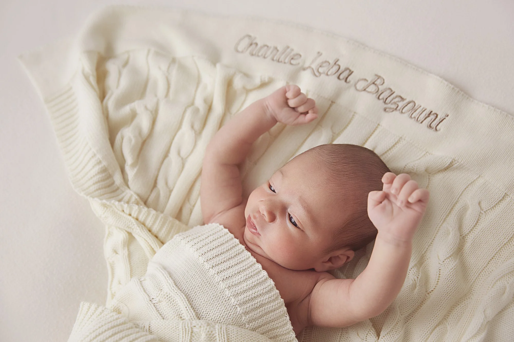 A newborn baby lying on a cream-colored knitted blanket with embroidered words and a pacifier pattern, wearing a matching cream knitted sweater and looking at its raised arms.