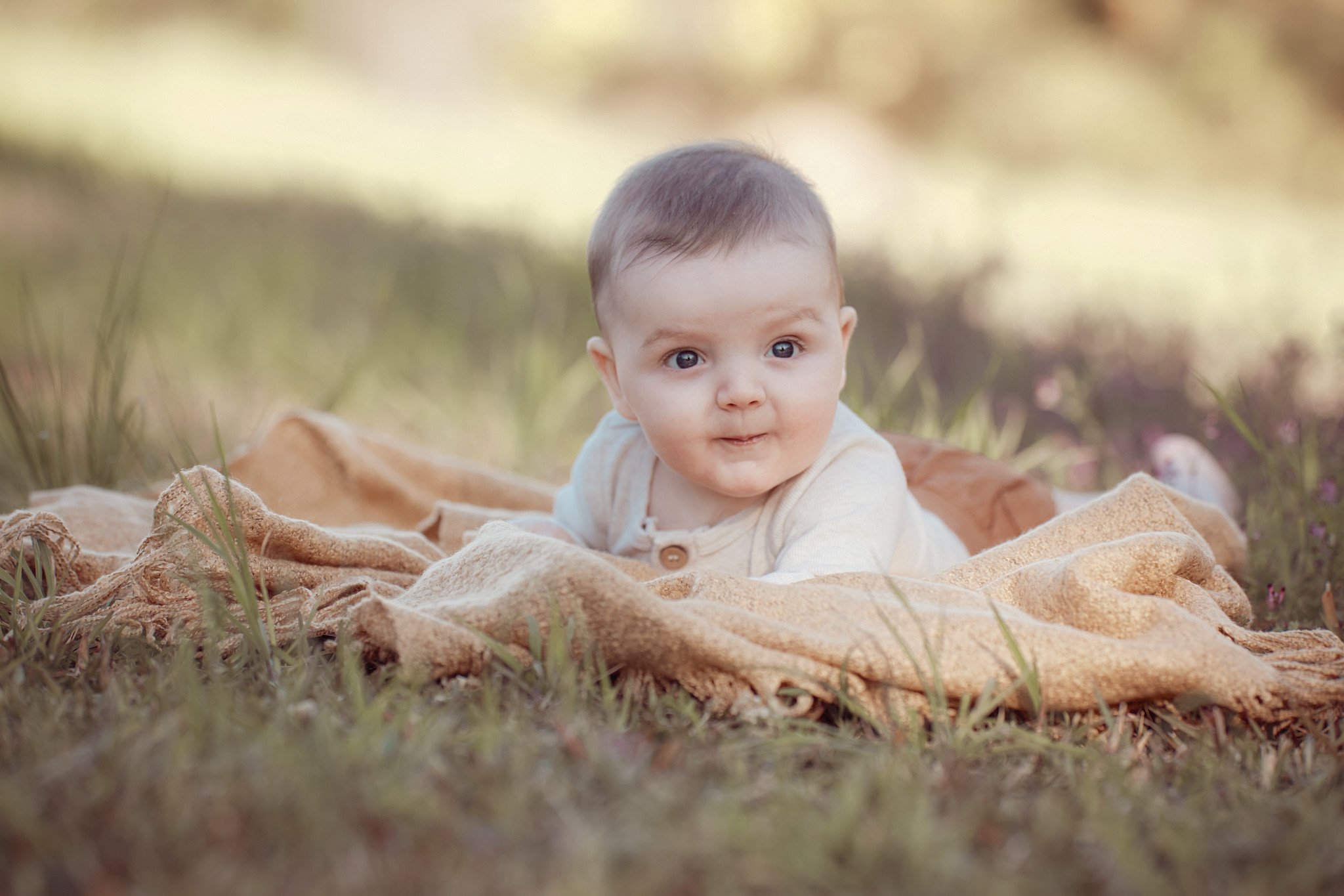 A baby lying on a blanket outdoors, surrounded by grass, with a blurred natural background.