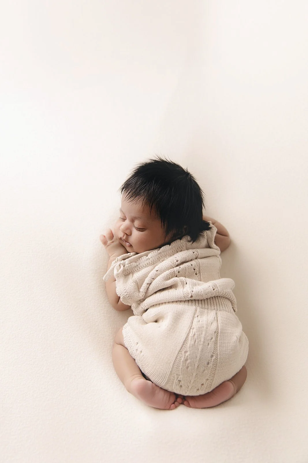 A sleeping baby with black hair, curled on a white surface, dressed in a beige knitted outfit, with eyes closed and a peaceful expression.