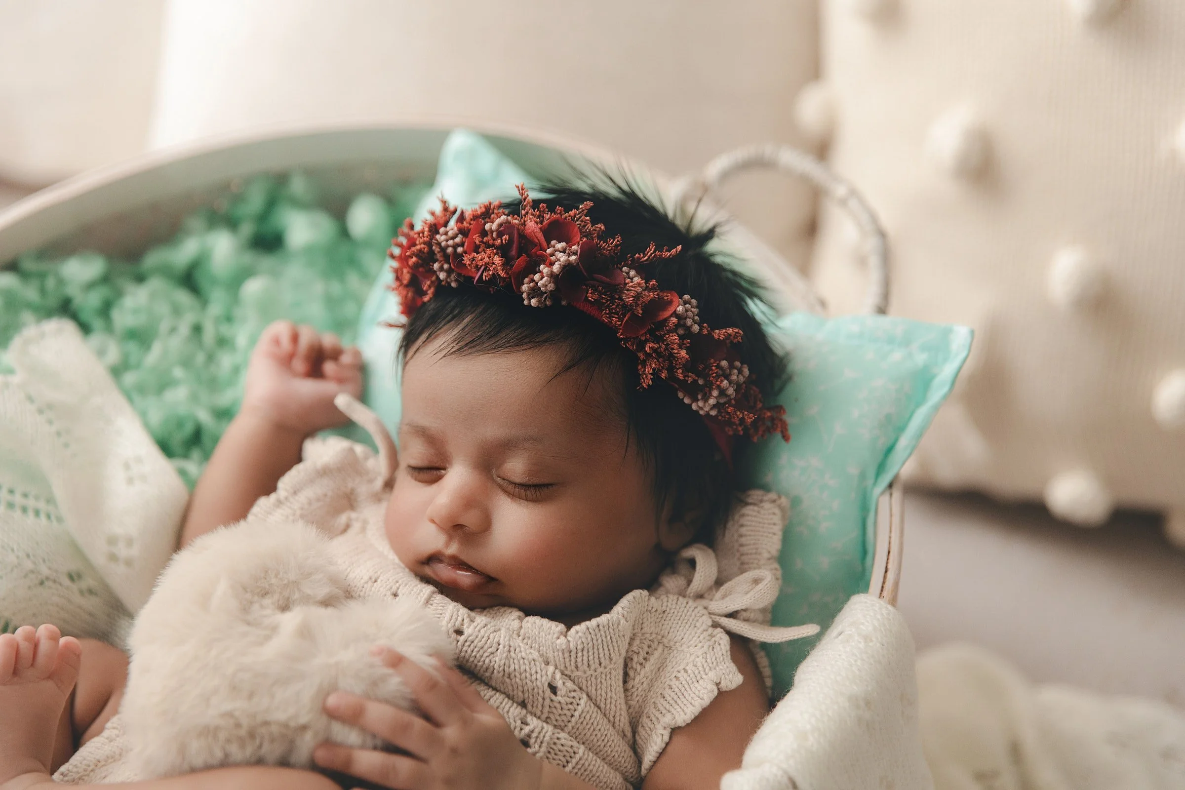 A newborn baby girl with short dark hair, wearing a beige knitted sweater, is sleeping in a hospital bed with a white fluffy animal. The baby has a floral headband with red flowers and small white beads on their head.