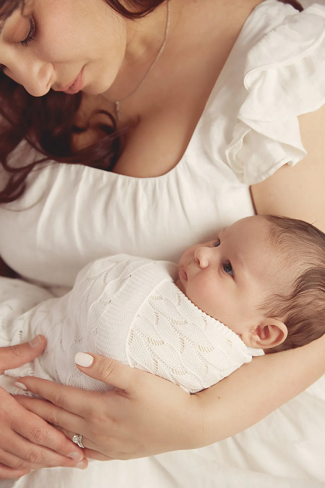A woman holding a baby close to her chest, both dressed in white. The woman is looking down at the baby, who is gazing up at her.