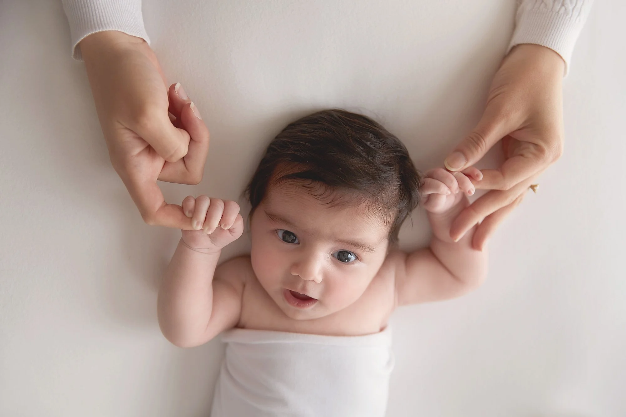 A baby with dark hair lying on a white surface, looking at the camera, holding hands with mum.