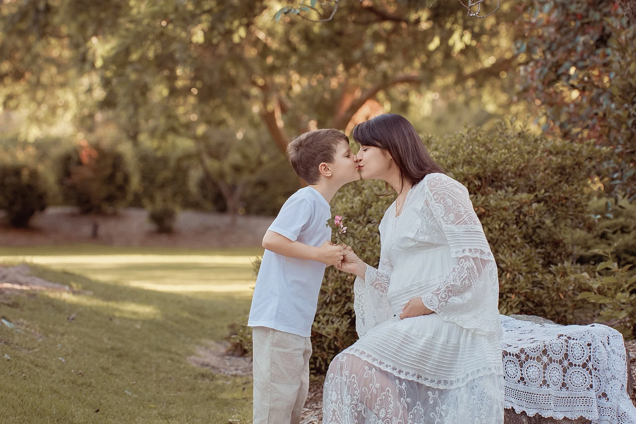 A woman and a young boy sharing a kiss outdoors in a park-like setting, with the woman sitting on a lace-covered bench and the boy holding a small flower.