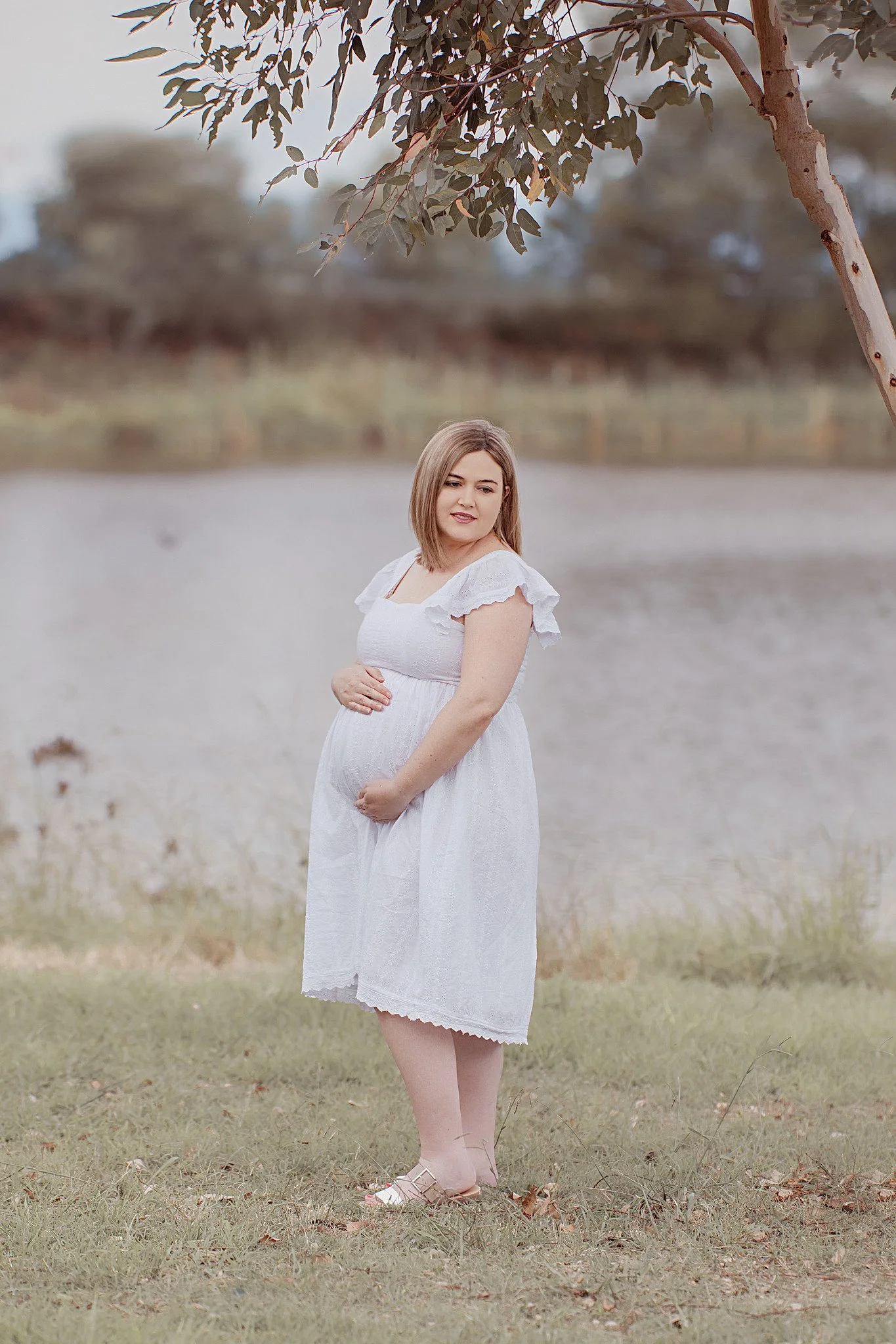 A pregnant woman in a white dress standing outdoors near a body of water, holding her belly, with trees and a cloudy sky in the background.
