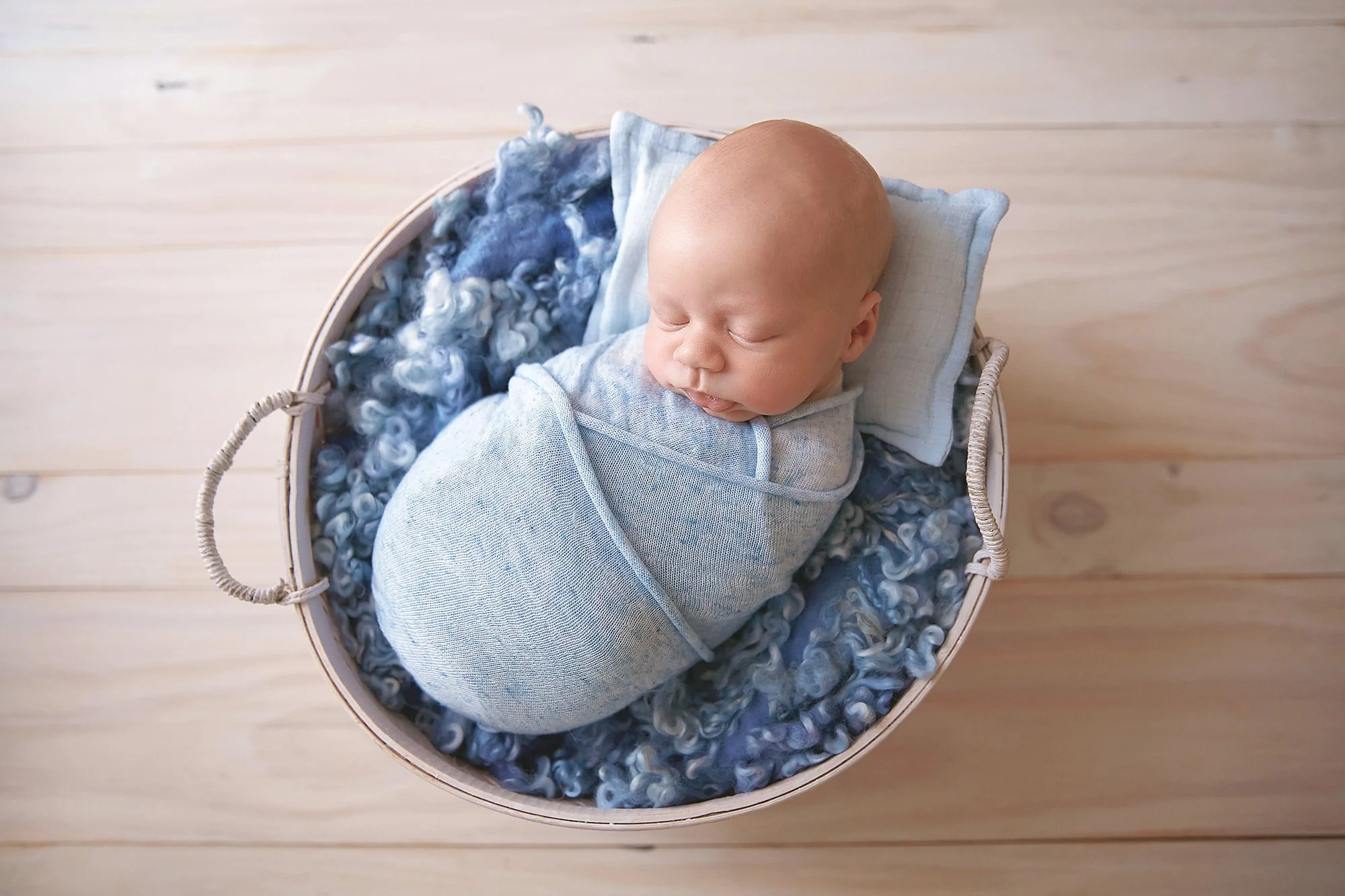 A baby is swaddled in a light blue blanket, sleeping in a round basket filled with blue and white curly wool and a small pillow against a light wooden floor.