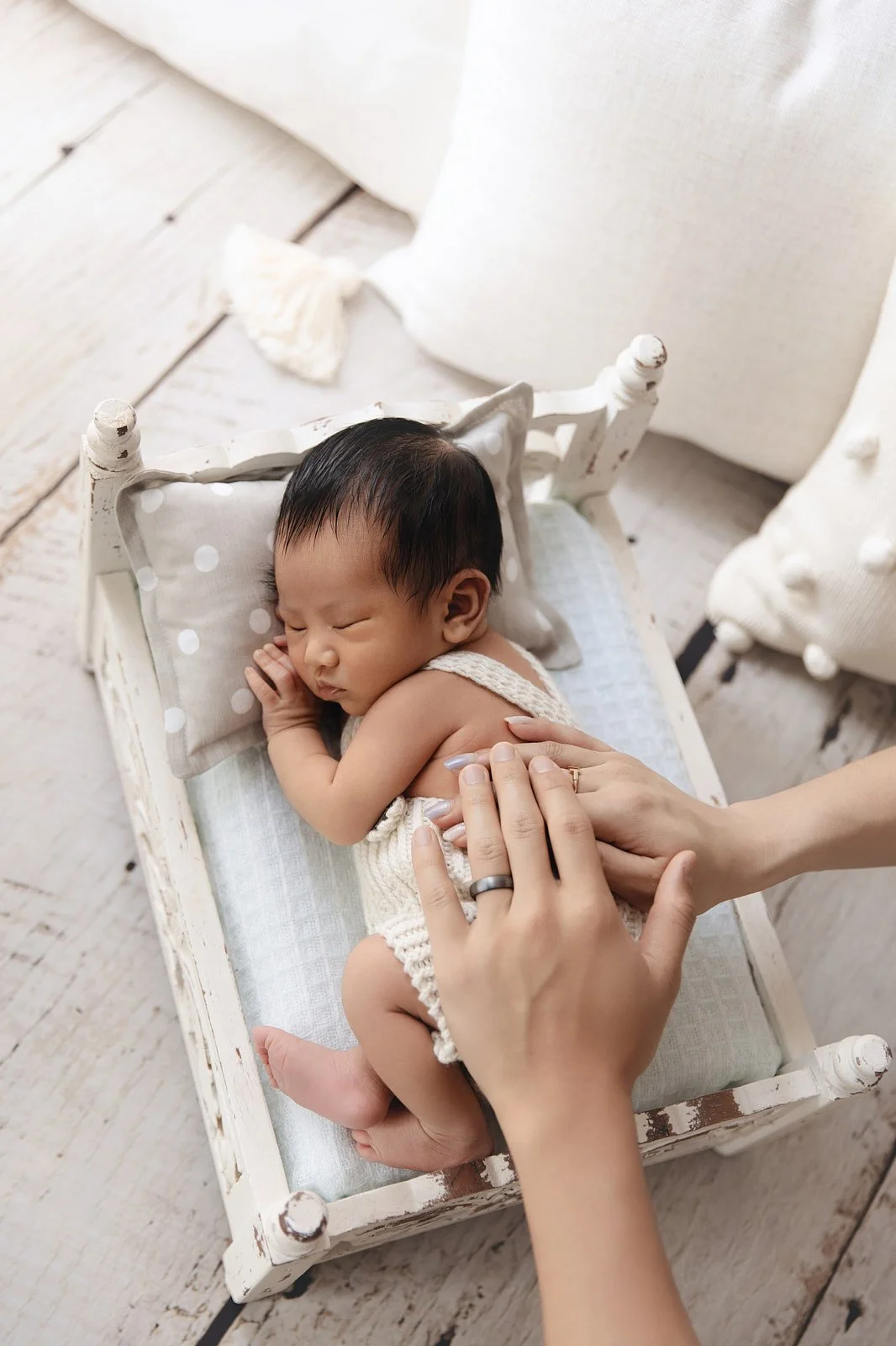 A sleeping baby in a small white wooden cradle with a pillow and blanket, being gently touched by an adult hand.