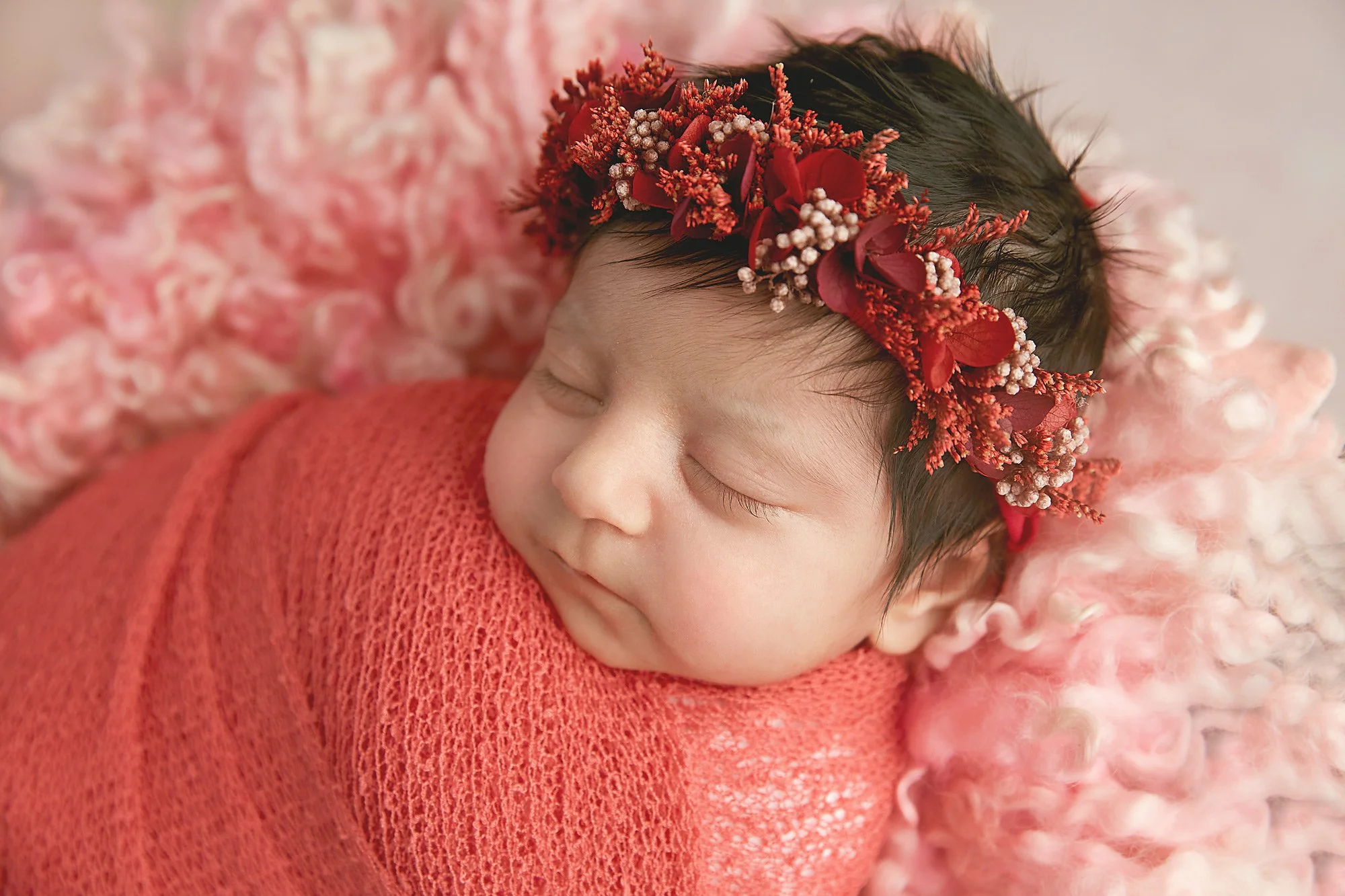 A sleeping baby girl with a floral headband, wrapped in a pink knitted blanket, lying on a soft pink fluffy surface.