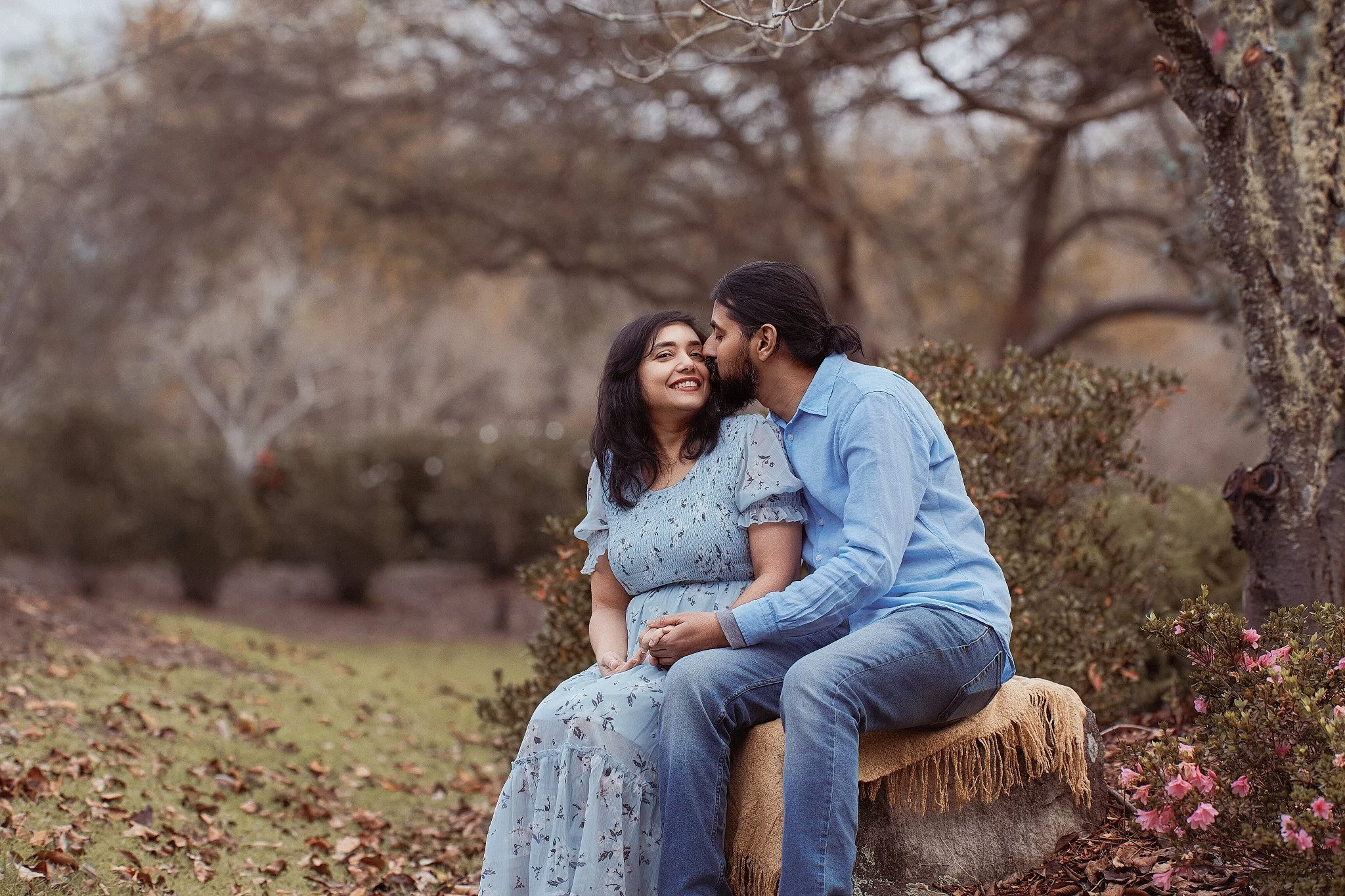 A couple sitting on a tree stump in a park with autumn trees, smiling. The man is kissing the woman's forehead, and she is smiling happily.