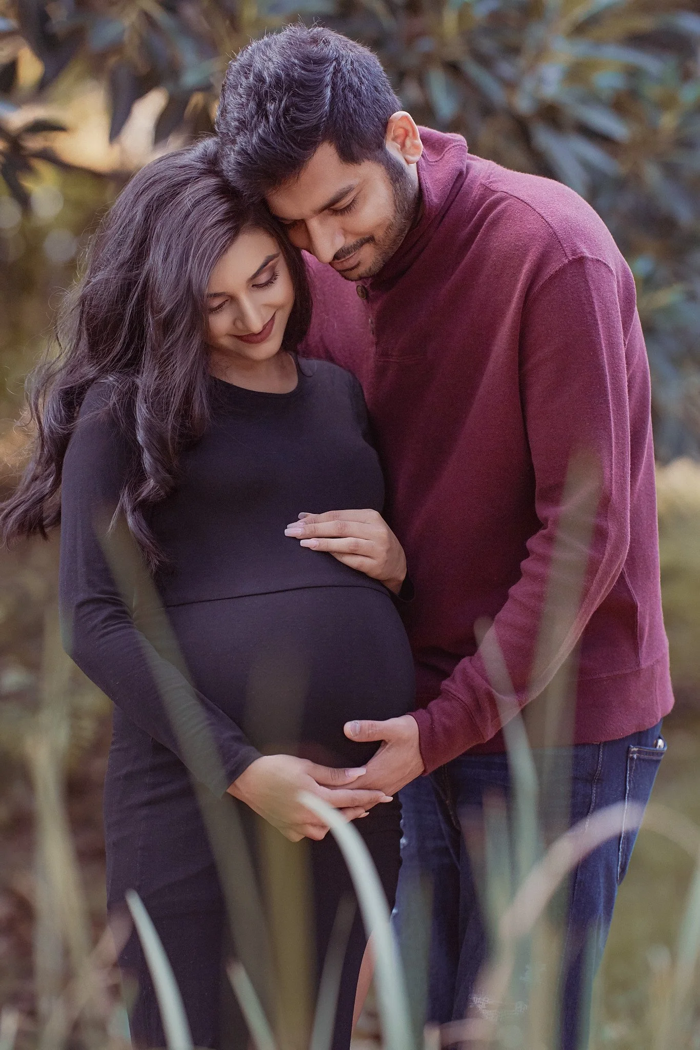 A pregnant woman and her partner standing outdoors, gently holding her belly, with both their hands on her stomach, creating a heart shape at their pregnancy shoot. They are smiling softly with their heads touching, surrounded by blurred greenery.