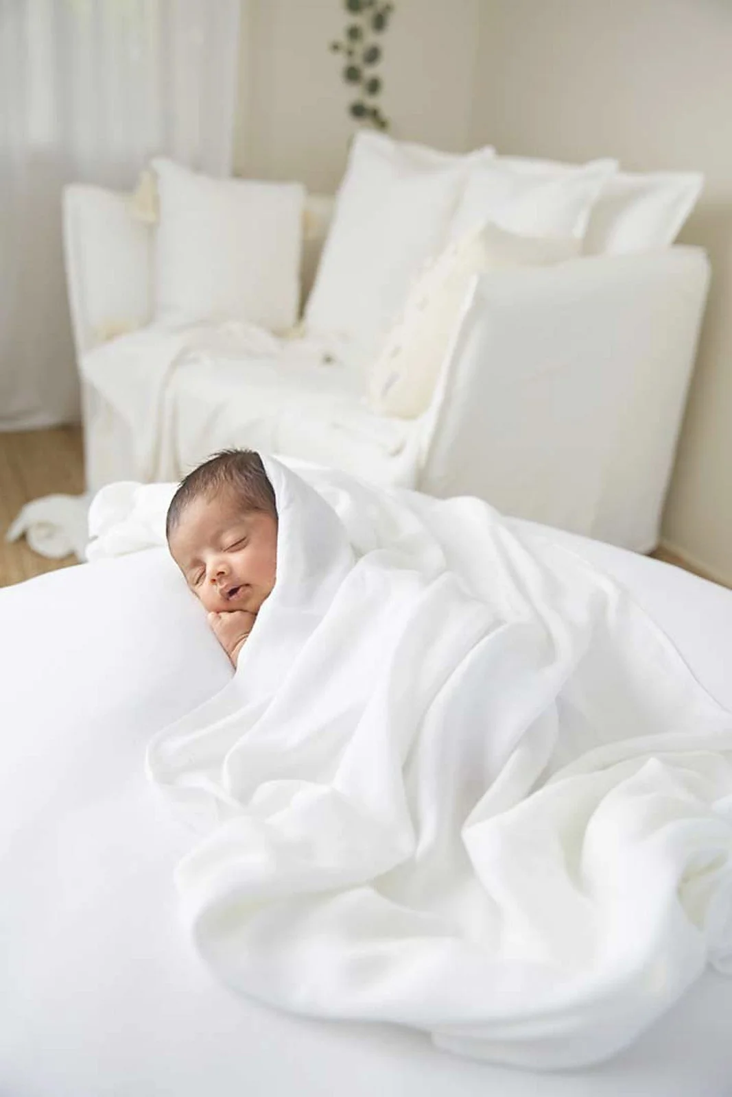 A baby sleeping on a white bed, wrapped in a white blanket, with pillows and a headboard in the background.