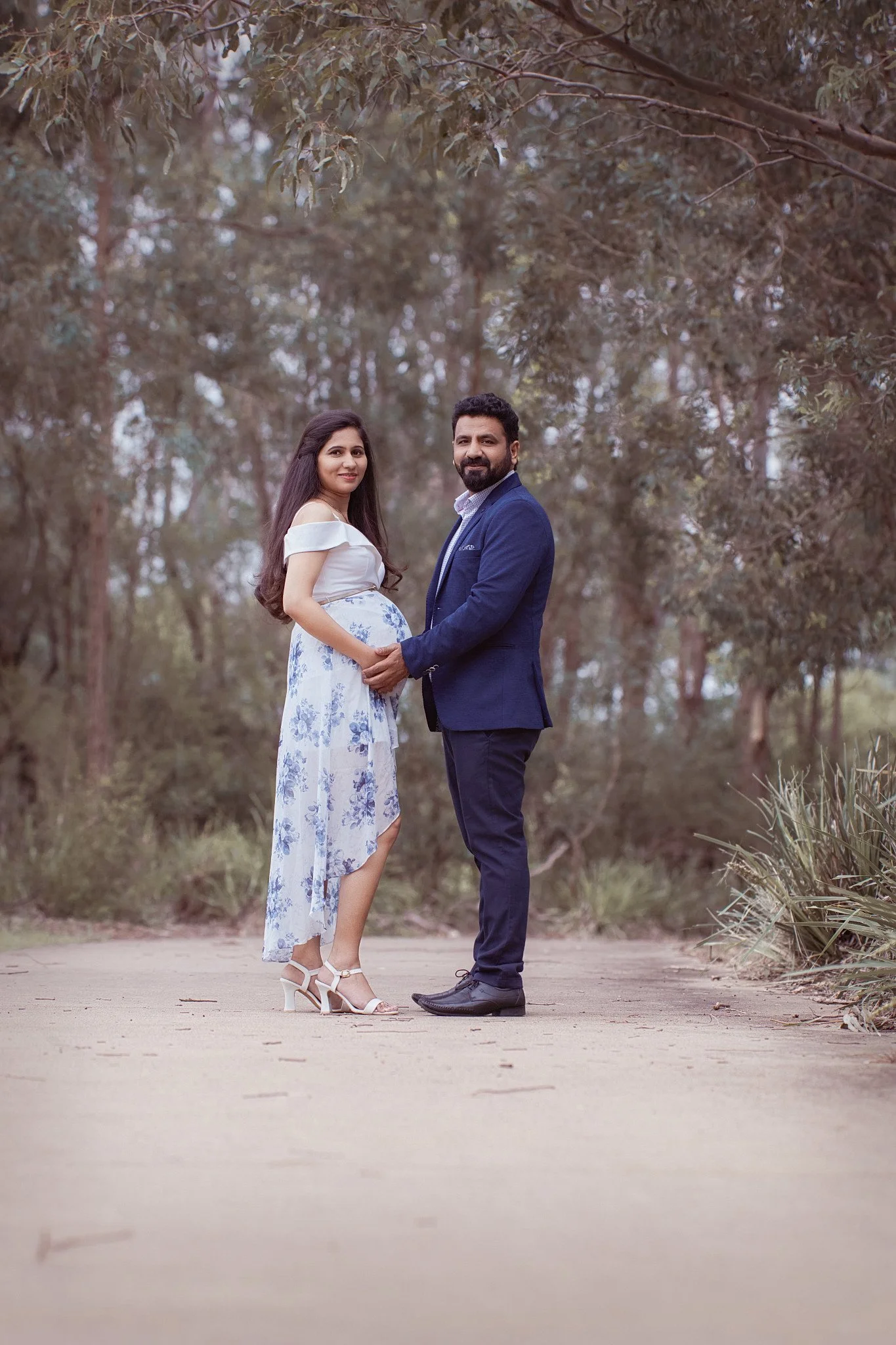 A pregnant woman and a man standing outdoors on a dirt path surrounded by trees, holding hands and smiling at the camera.
