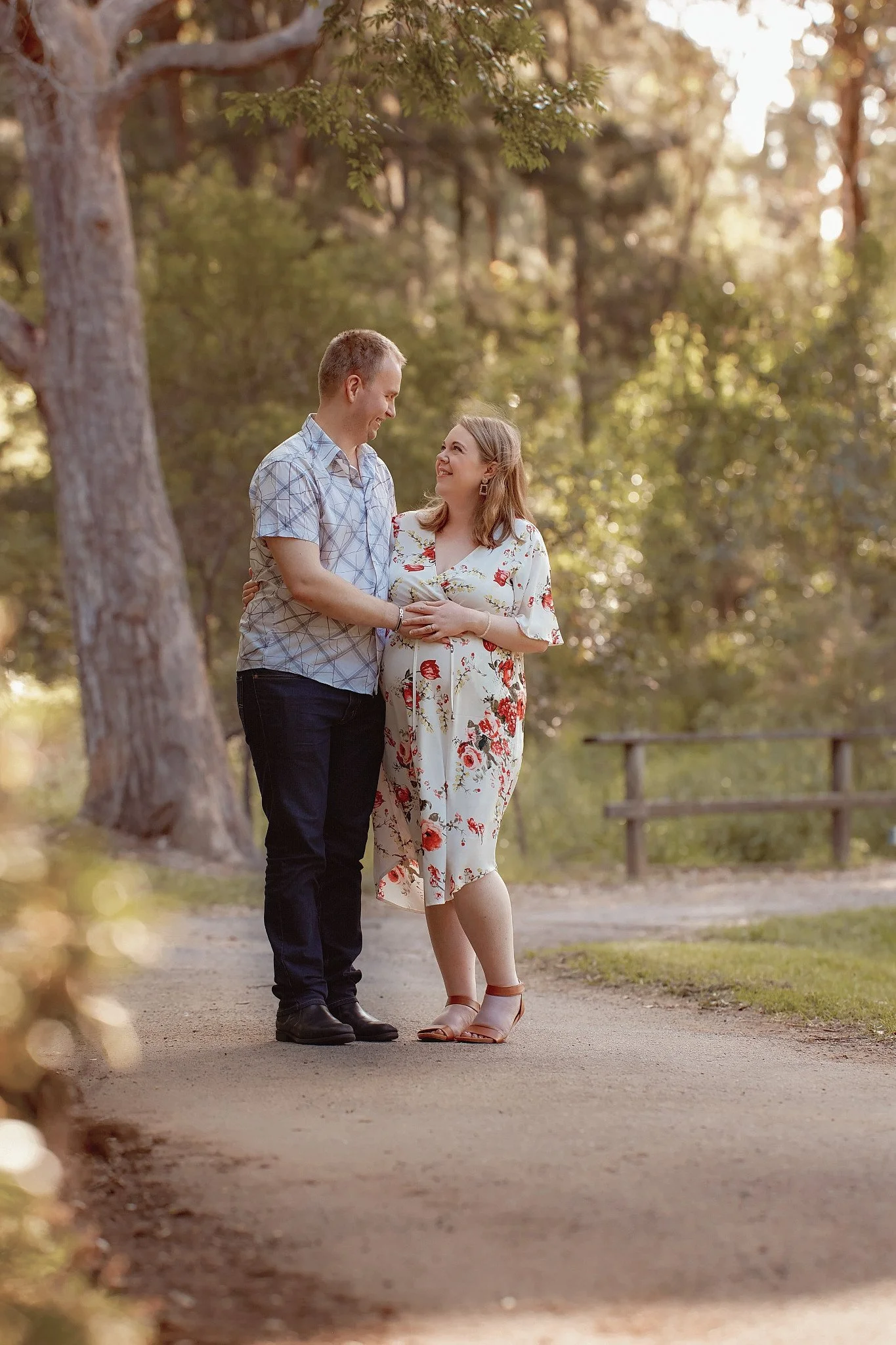 A couple standing closely together on a park path, smiling and looking into each other's eyes, surrounded by trees and warm sunlight.
