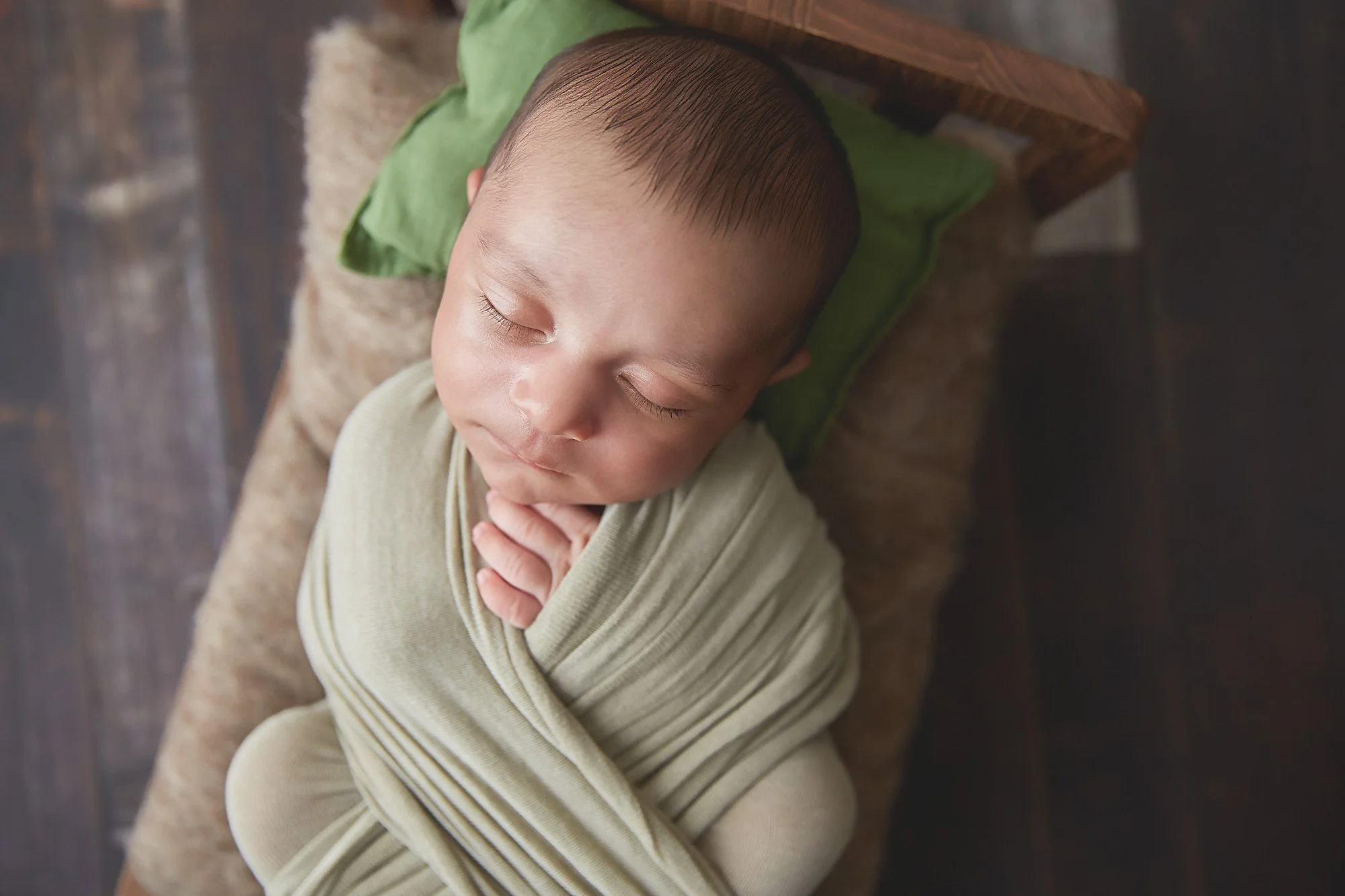 A young child with closed eyes, resting their head on a green pillow and wrapped in a beige blanket, lying on a plush brown cushion on a wooden floor.