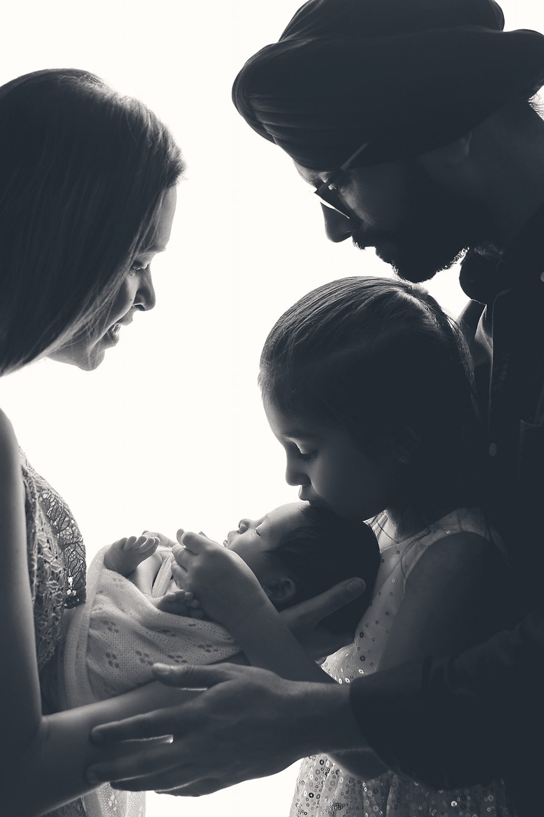 A family holding their newborn baby, with the mother and father looking down at the infant and the older sister gently touching the baby's face, in black and white.
