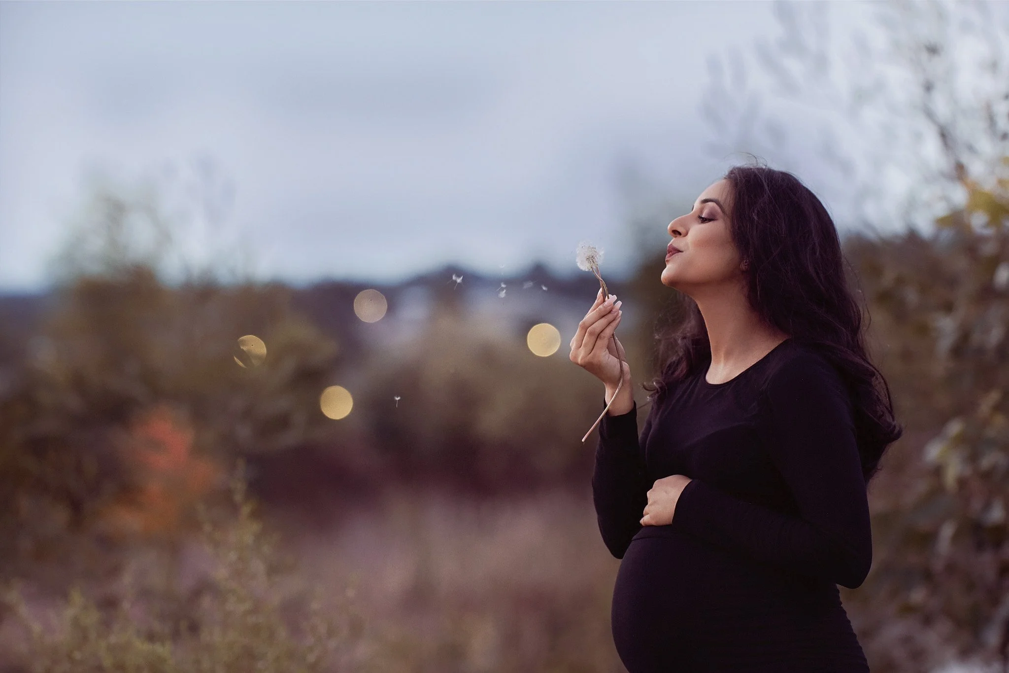 Pregnant woman outdoors holding a dandelion with seeds floating away, blurry background with trees and bokeh lights.