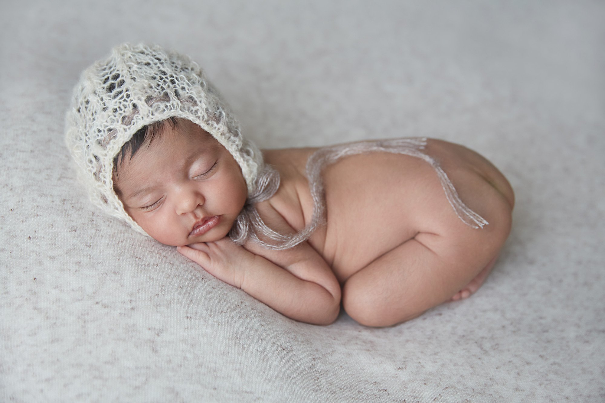 A sleeping newborn baby with a knitted hat, lying on a soft surface, in a fetal position with hands under the cheek.