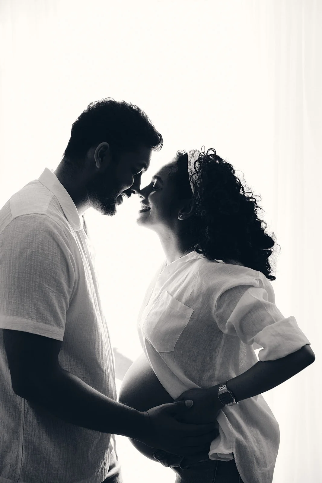 Black and white photo of a smiling pregnant woman and a man, facing each other with their foreheads touching, holding hands, backlit with natural light.