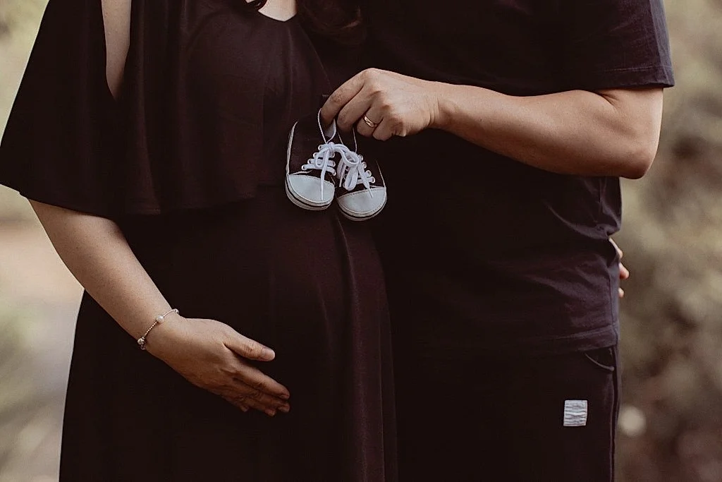 A pregnant woman is holding a pair of baby shoes, with one hand on her belly and another hand holding the shoes, alongside a man who has one arm around her.