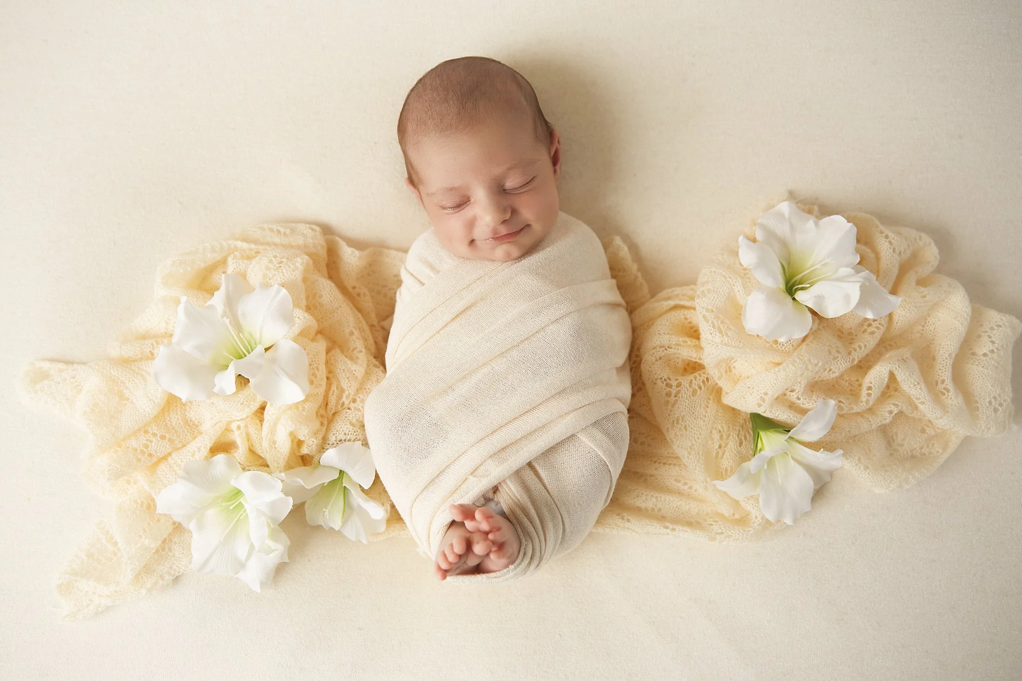 A newborn baby wrapped in a light cream swaddle, sleeping on a cream blanket, surrounded by lace and white flowers.