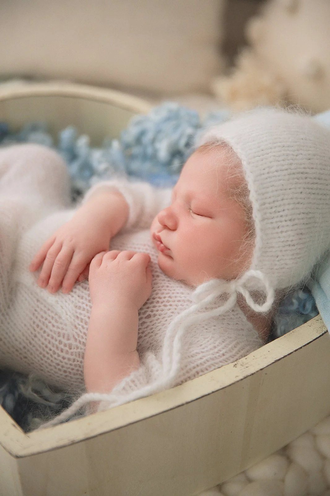 A sleeping baby with a knitted hat and sweater lying in a box, surrounded by soft blue and white blankets.