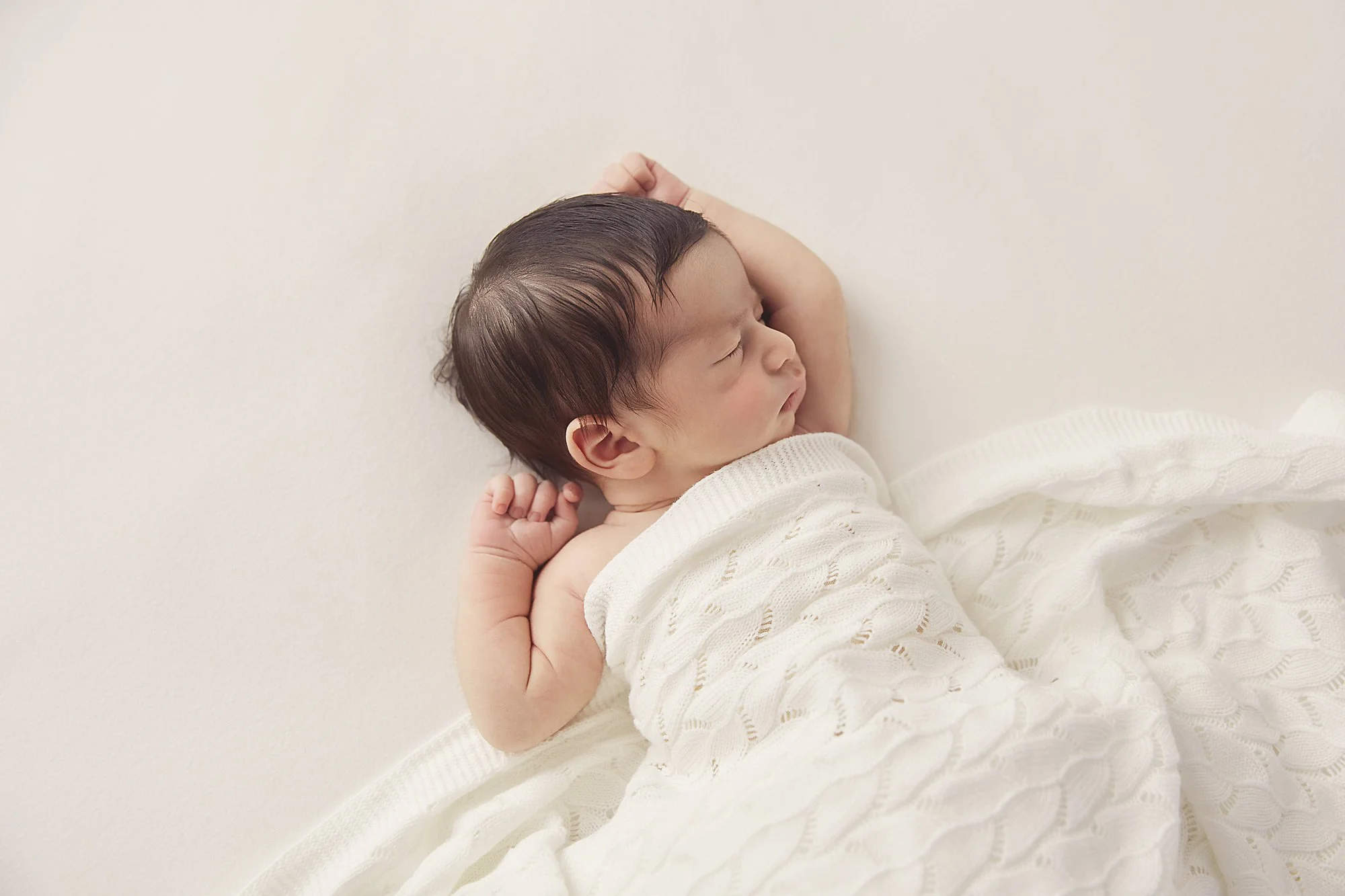A sleeping baby with dark hair lying on a white blanket with a plain white wall background.