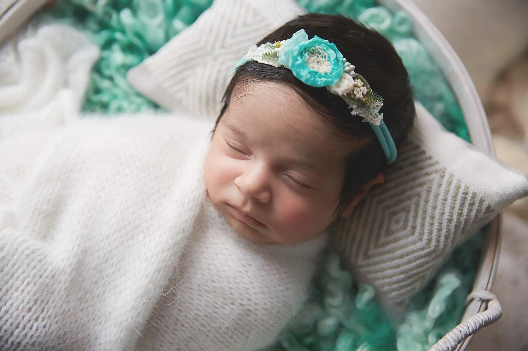 A sleeping baby girl with a flower headband, wrapped in a white blanket, resting on a decorative pillow in a wicker basket.