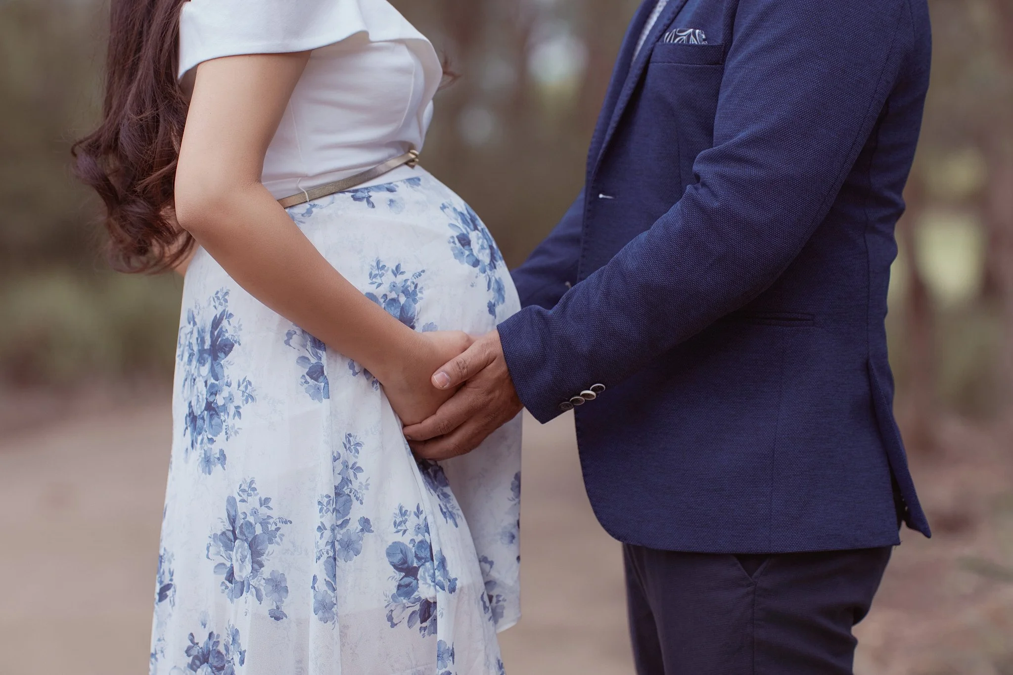 A couple holding hands, with the woman wearing a white dress with blue floral patterns and the man in a navy blue suit, standing outdoors.